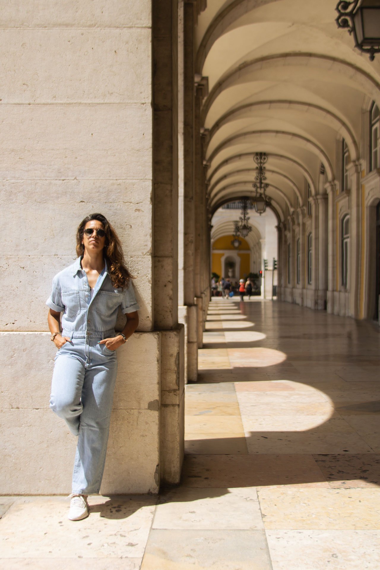 Mulher com cabelos castanhos, óculos escuros, vestindo macacão jeans, apoiada na parede de um prédio antigo com arcos, sob sombra e ao sol, em uma área de passeio urbano.
