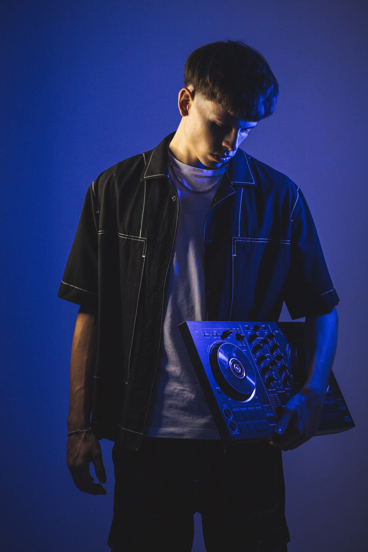 Jovem DJ segurando um mixer de música em um fundo azul escuro, vestindo uma camiseta branca e uma jaqueta preta com costuras brancas.