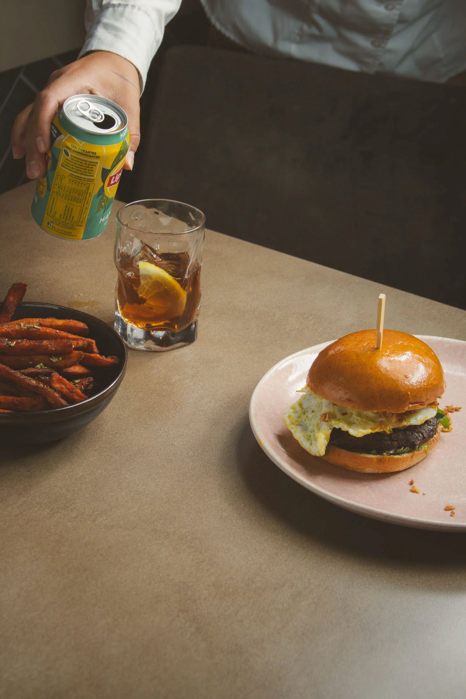 Hambúrguer com ovo frito, batatas fritas temperadas, lata de refrigerante e copo de refrigerante com gelo e limão sobre mesa