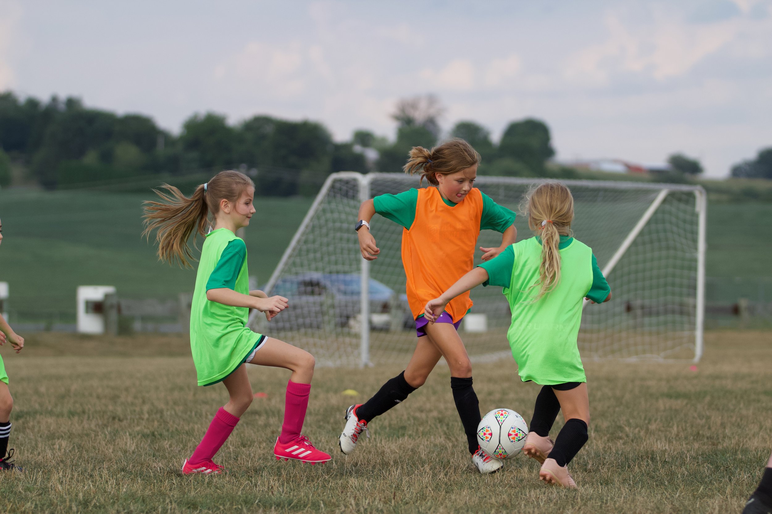 Young girls playing soccer on a grassy field, wearing bright green and orange jerseys, with a goal in the background.