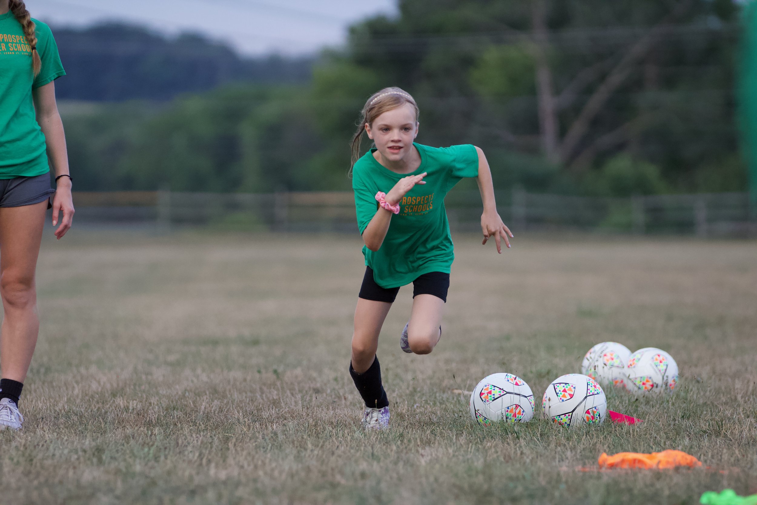 A young girl in a green shirt running on a soccer field with multiple soccer balls visible on the grass.