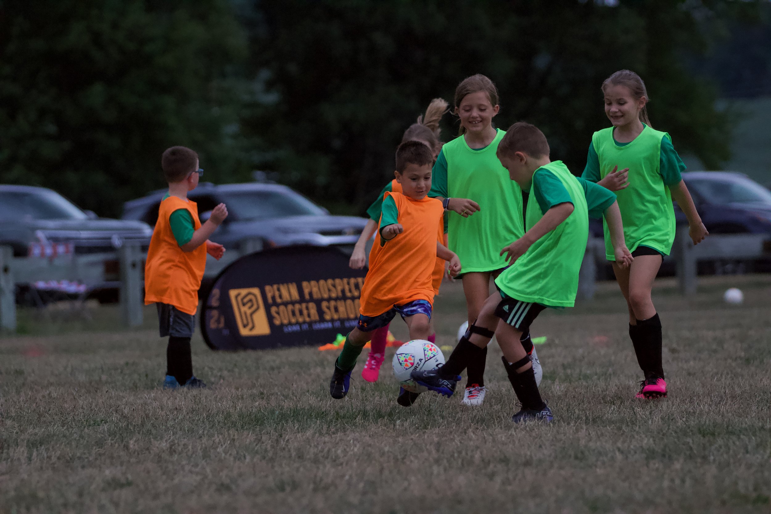 Children playing soccer on a field, wearing green and orange jerseys, with a "Penn Prospects Soccer School" sign in the background.