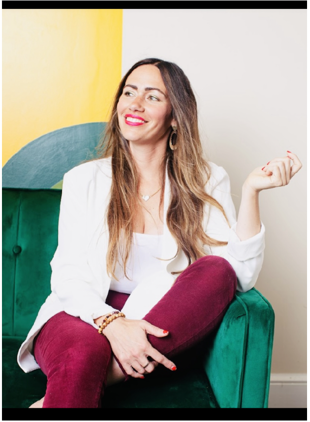 Woman sitting on a green couch with a colorful yellow and blue wall behind her, smiling and looking to the side.