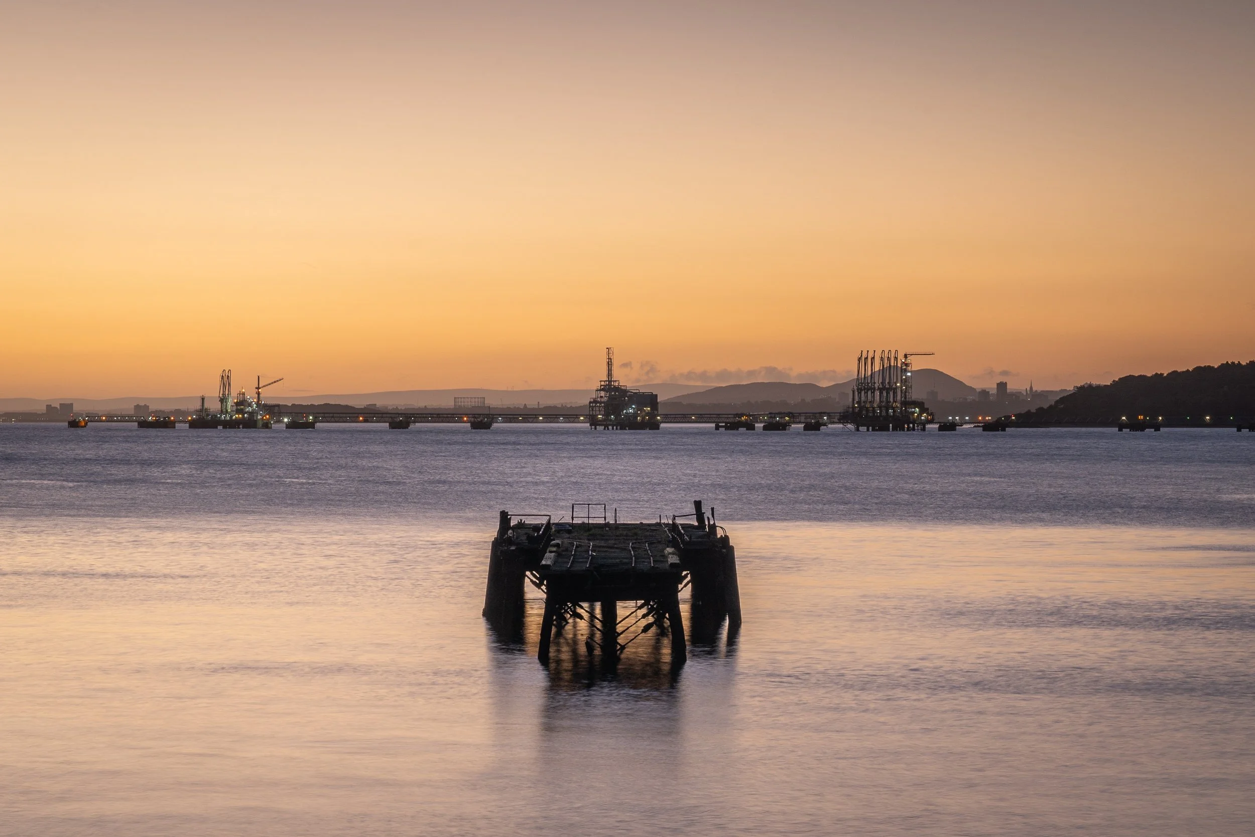 A sunset over a calm body of water with an old, rusted dock in the foreground and industrial structures, cranes, and distant hills on the horizon.