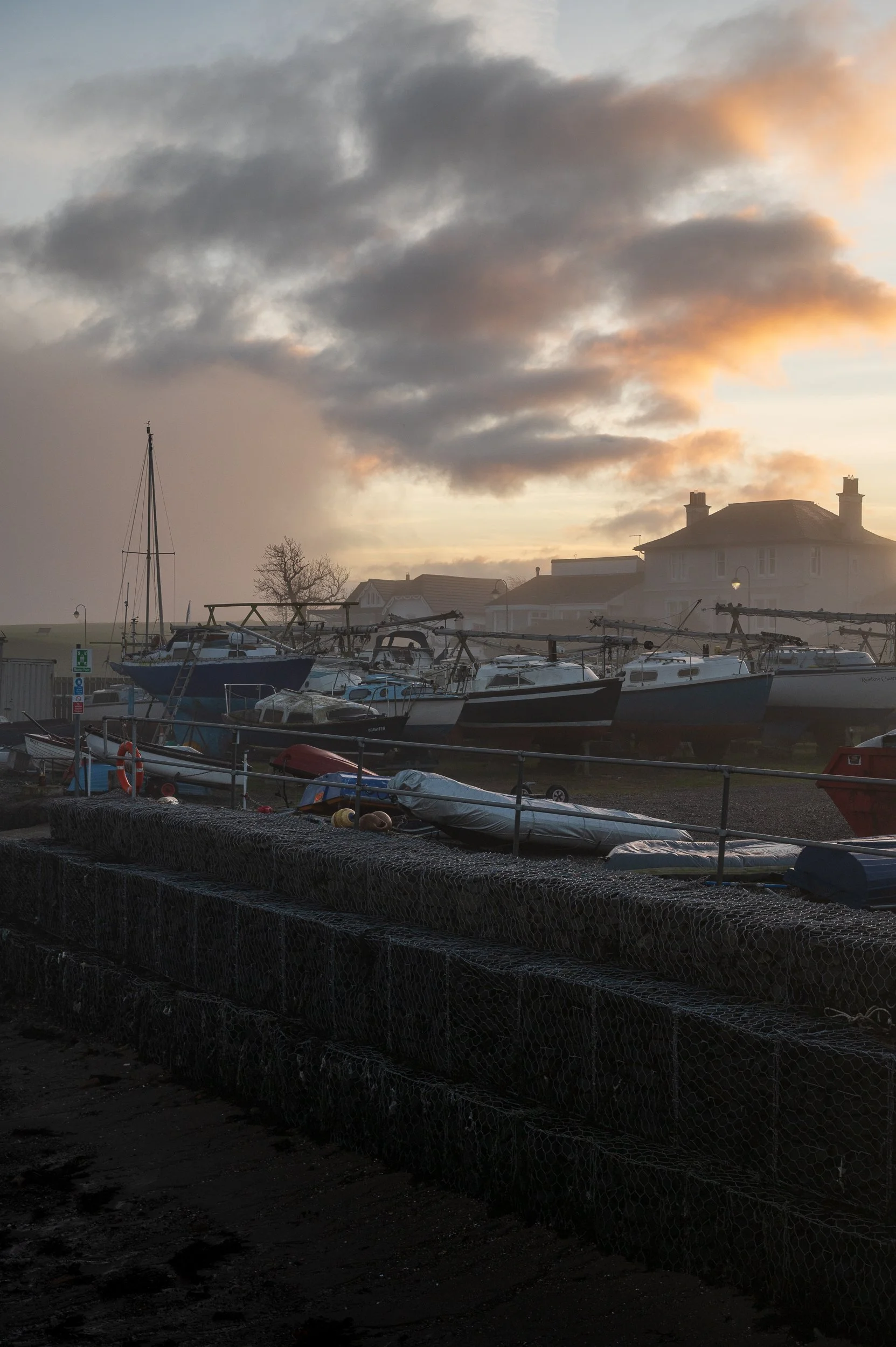A marina during sunset or early morning with several boats docked, some on land, under a cloudy sky with orange hues.