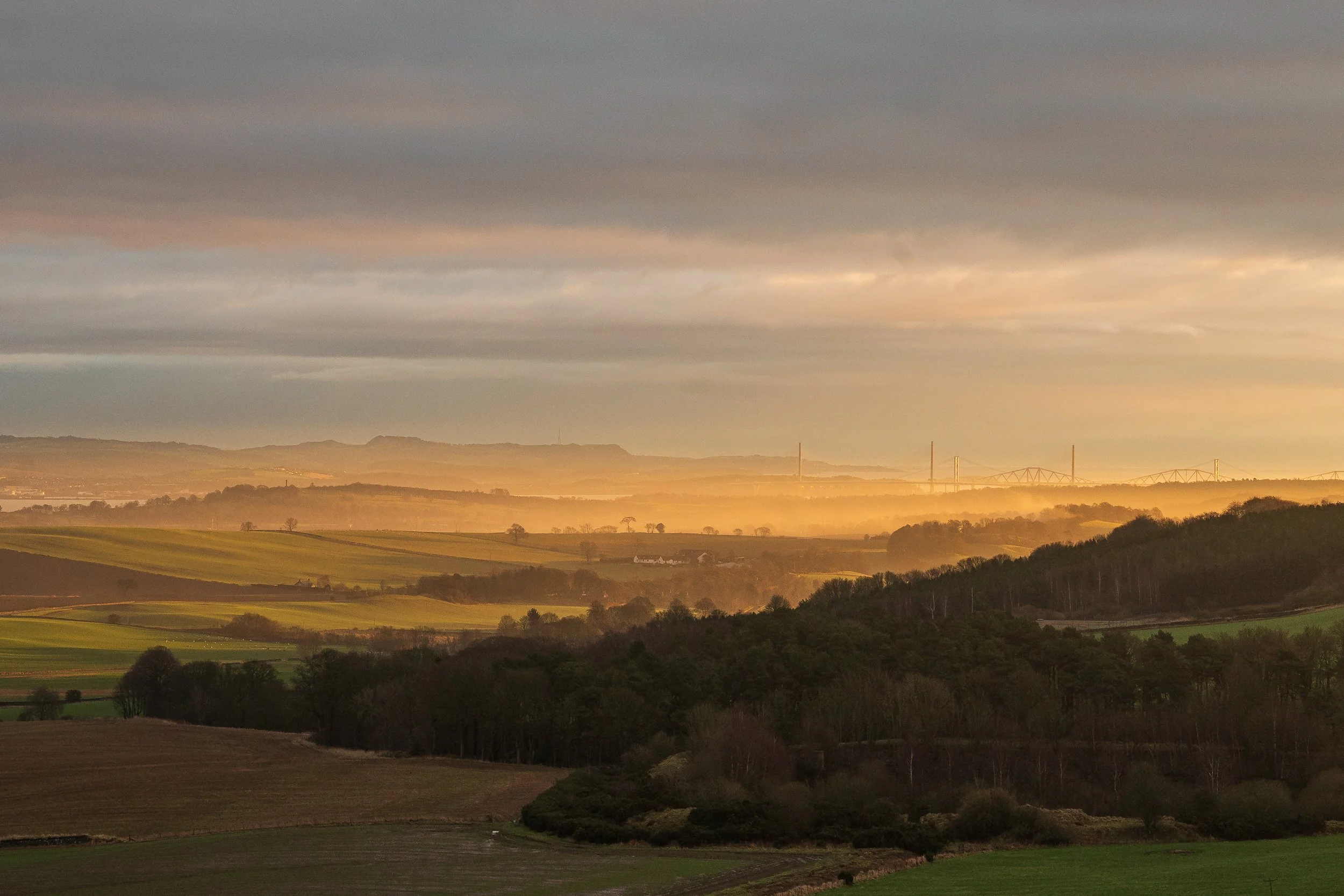 A scenic landscape of rolling hills and fields during sunset or sunrise, with a cloudy sky and a distant bridge on the horizon.