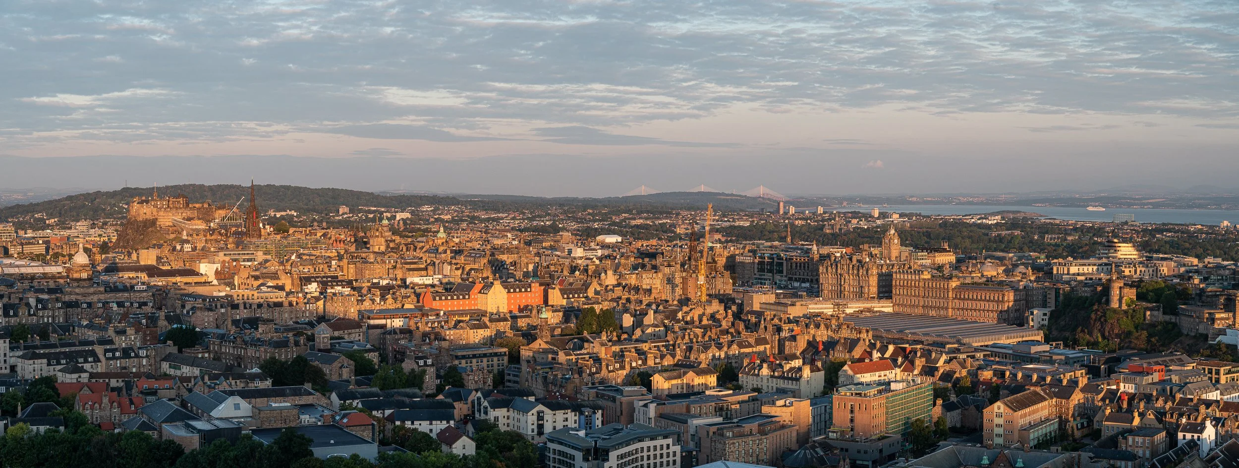 A cityscape in the evening with historic and modern buildings, a hilltop castle in the background, and a large body of water in the far distance.