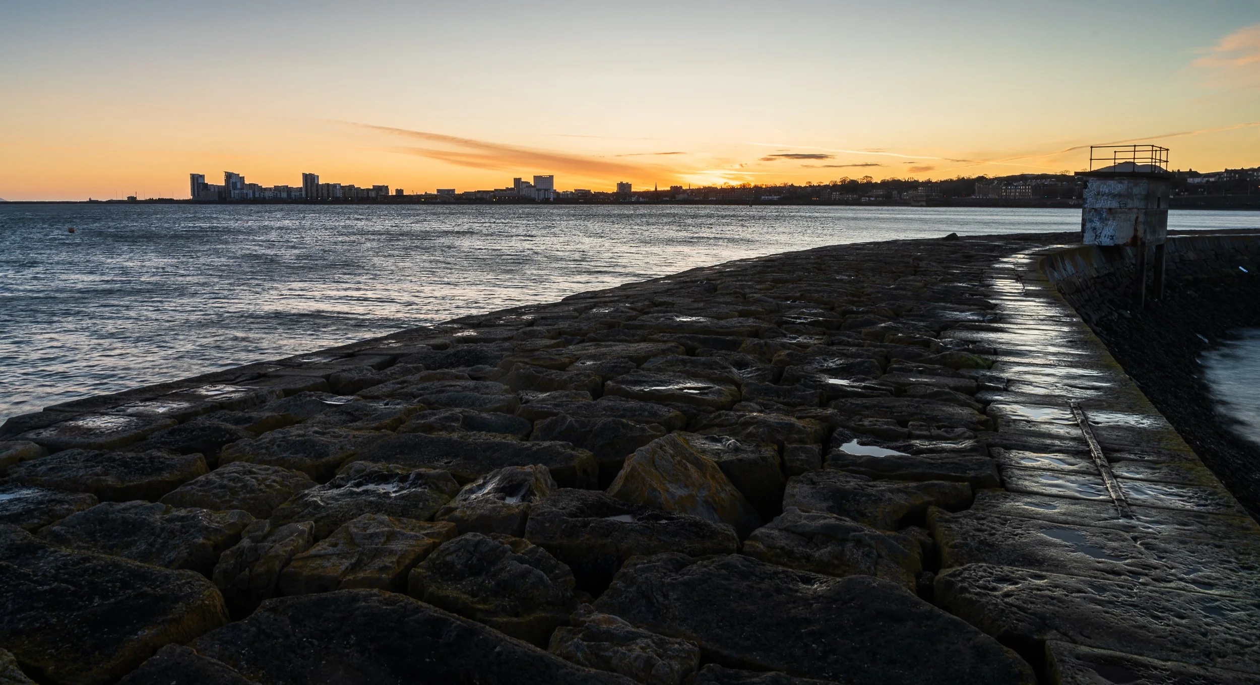 Sunset over a rocky waterfront with a distant city skyline in the background.