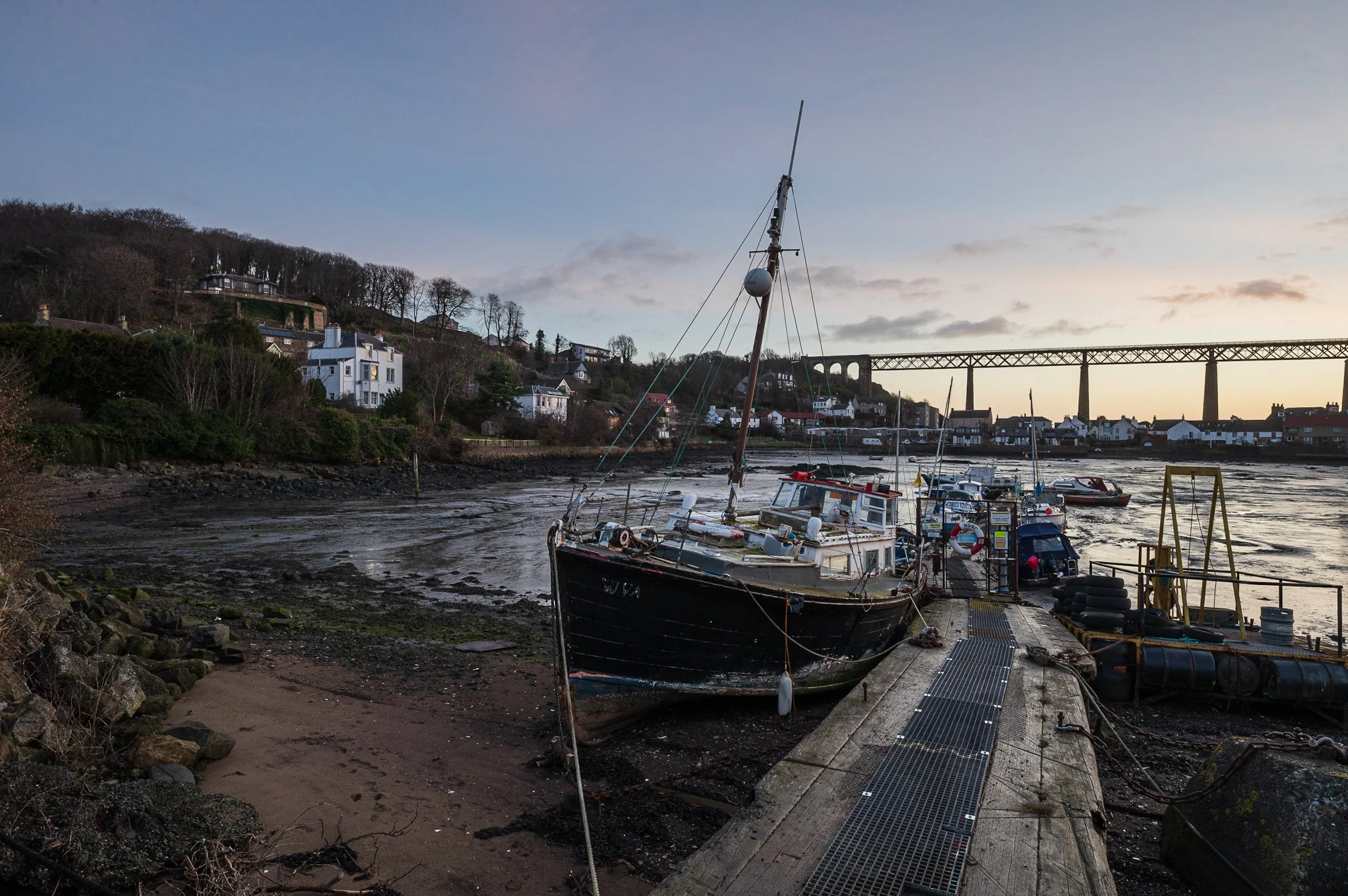 Boats docked at a pier on a cloudy evening with a hilly coastal town and train bridge in the background.