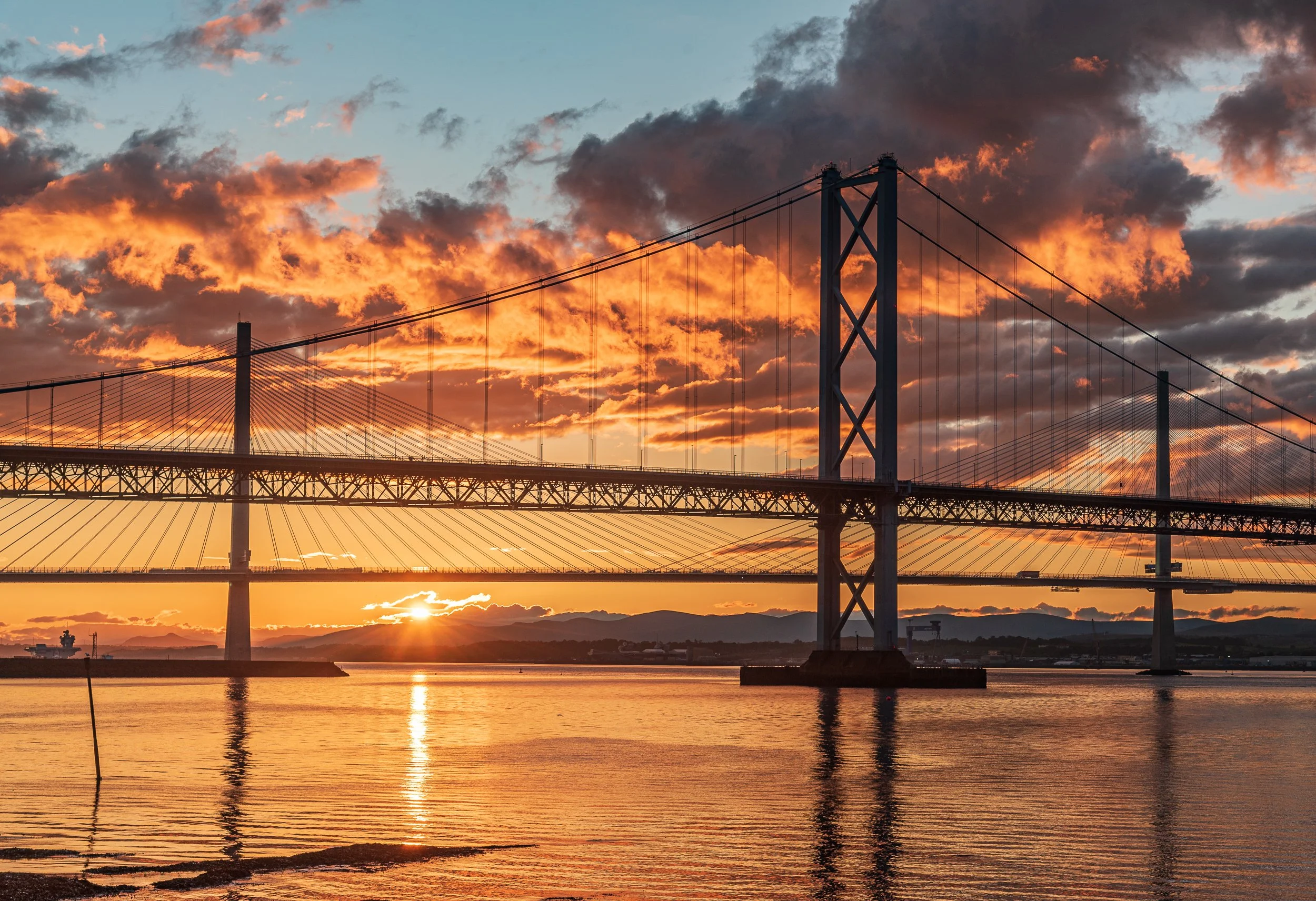 Sunset over a large suspension bridge spanning across a body of water, with orange and dark clouds in the sky.