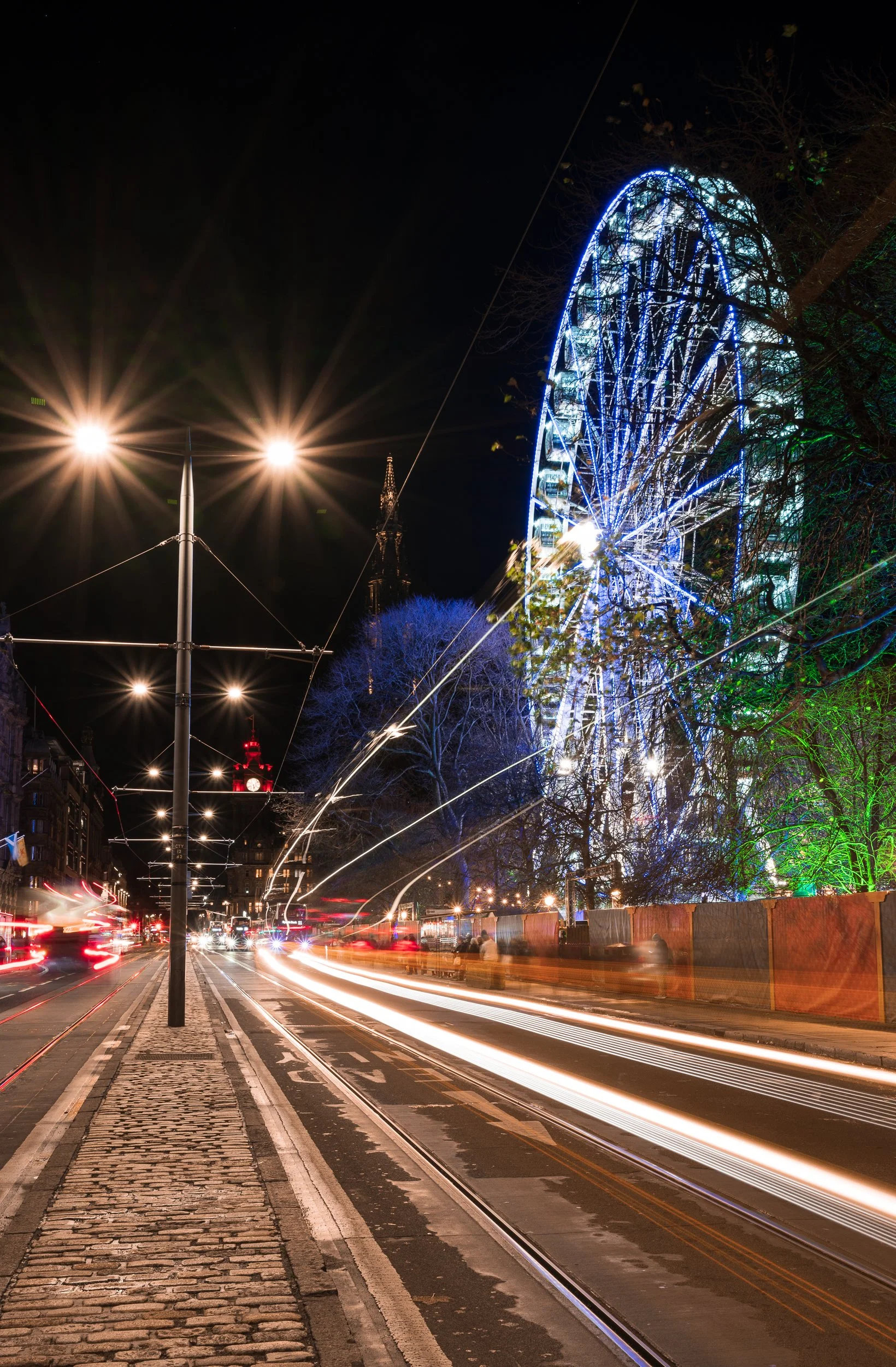 Nighttime city street with streaks of light from moving vehicles, illuminated Ferris wheel, streetlights, a red clock tower, and decorated trees.
