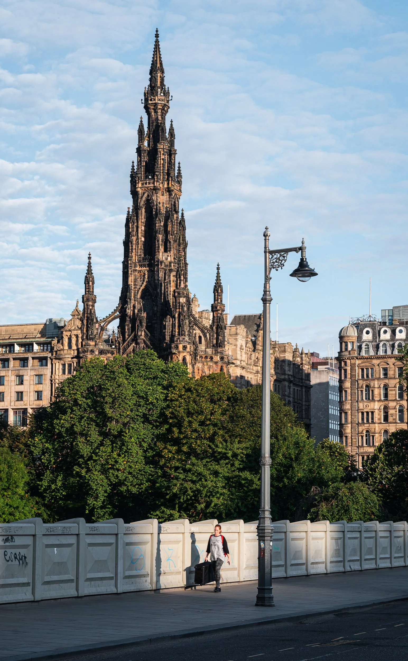 A person walking along a city street with a rolling suitcase, in front of a fence and trees, with a tall Gothic-style church or cathedral, and older buildings in the background, under a blue sky with scattered clouds.