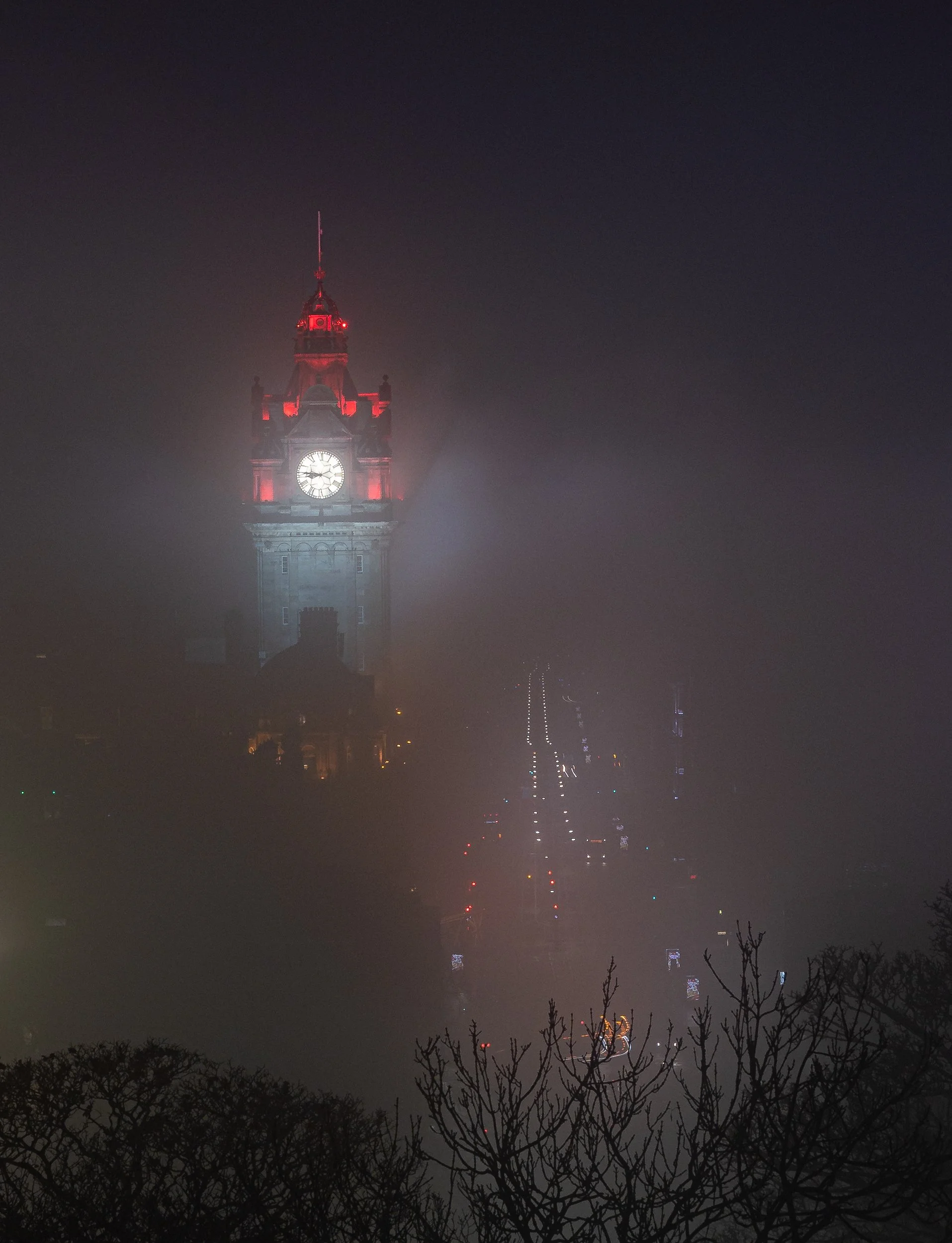 Nighttime view of a large clock tower shrouded in fog, with a faint city street and trees in the foreground.