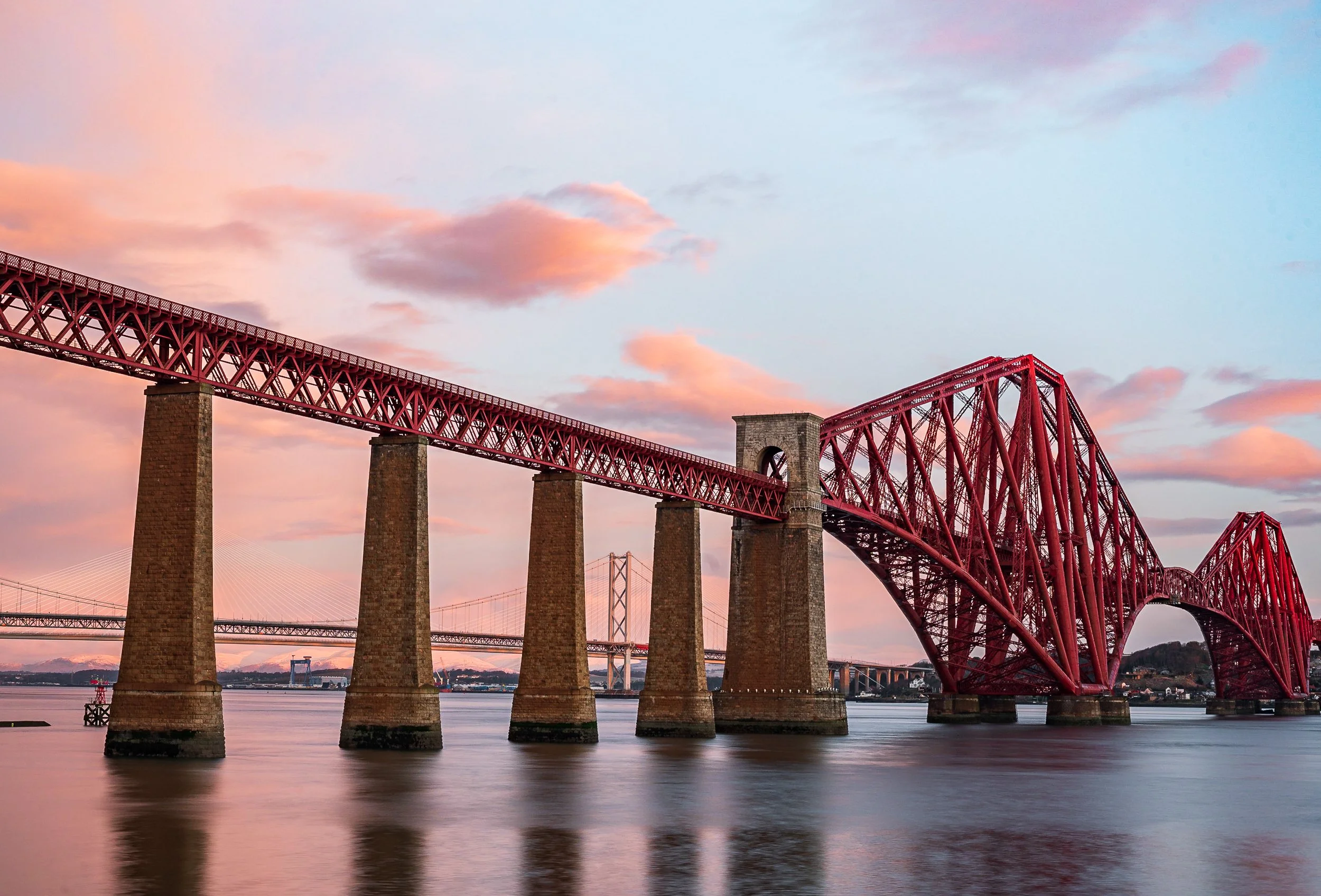 The image shows the Forth Bridge, a red cantilever railway bridge over the Firth of Forth in Scotland, with a sunset sky in the background and calm water below.