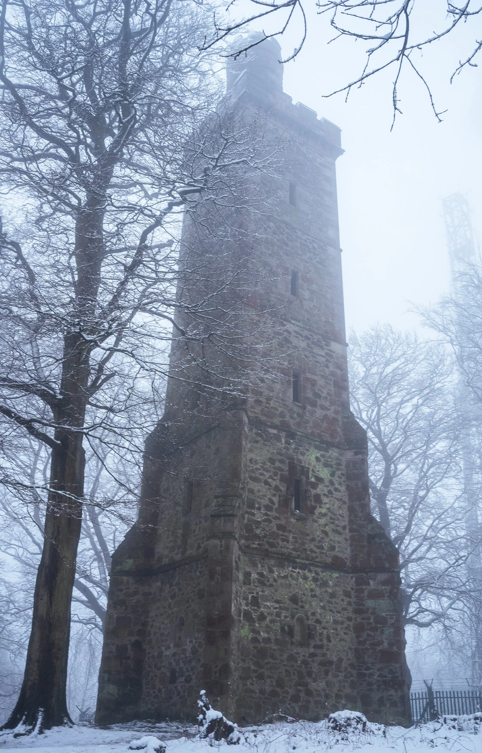 A tall, old stone tower surrounded by leafless trees with snow on the ground and foggy weather.