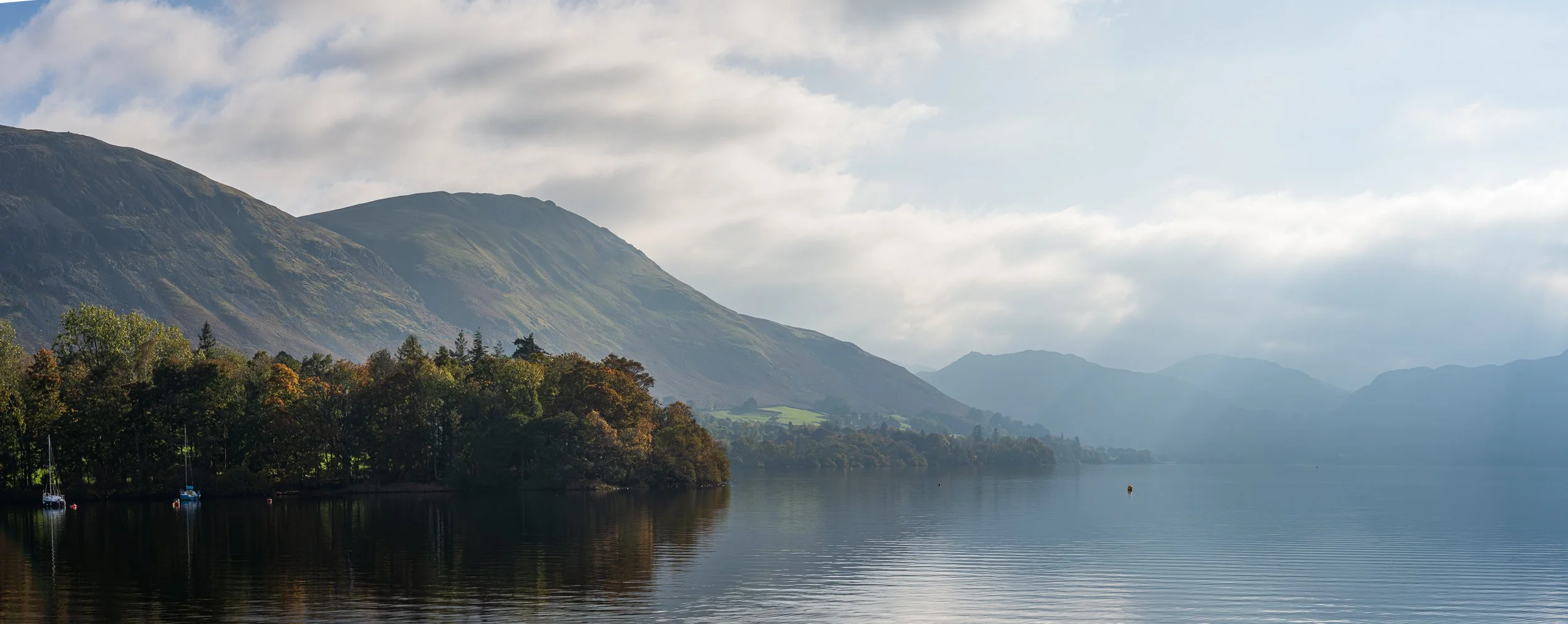 A peaceful lake surrounded by green trees with sailboats, mountains in the background, and partly cloudy sky.