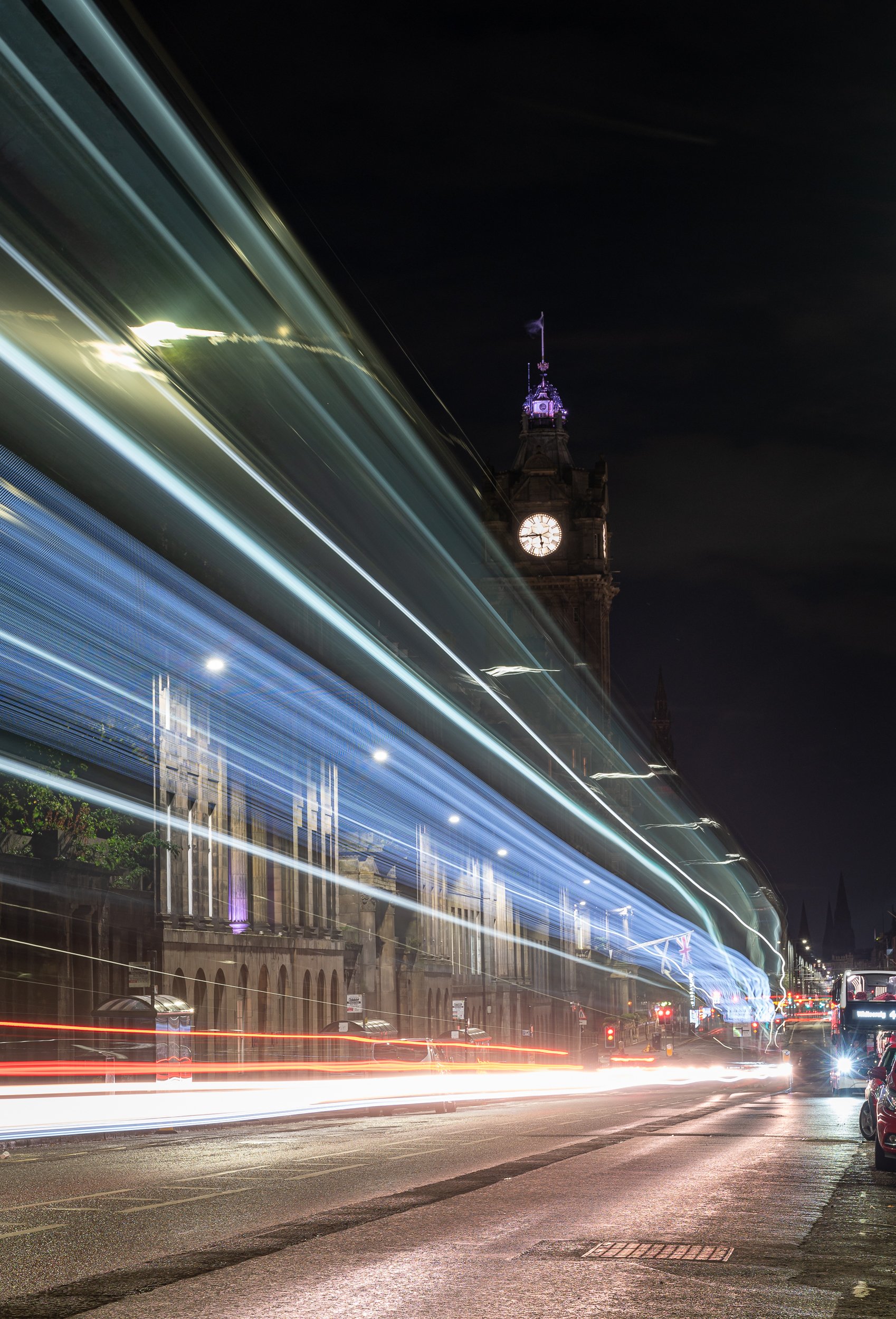 Nighttime city street scene with light trails from moving vehicles, and a historic clock tower in the background.