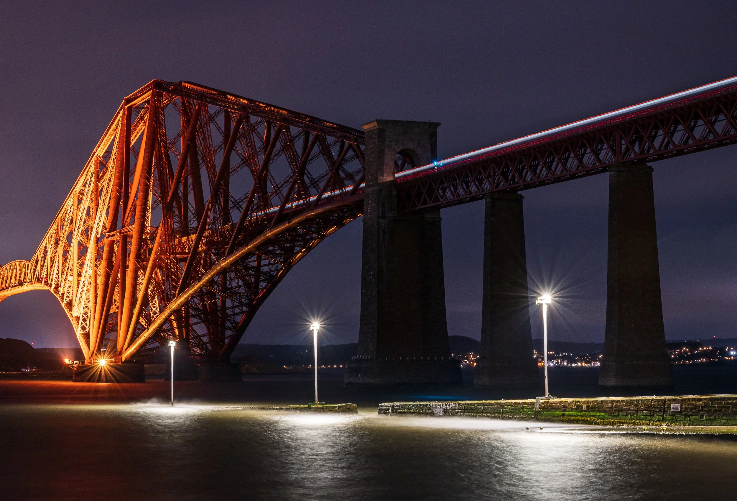 Night view of a large red steel bridge illuminated with orange lights, spanning over water with a city in the background, and streetlights illuminating the shoreline.