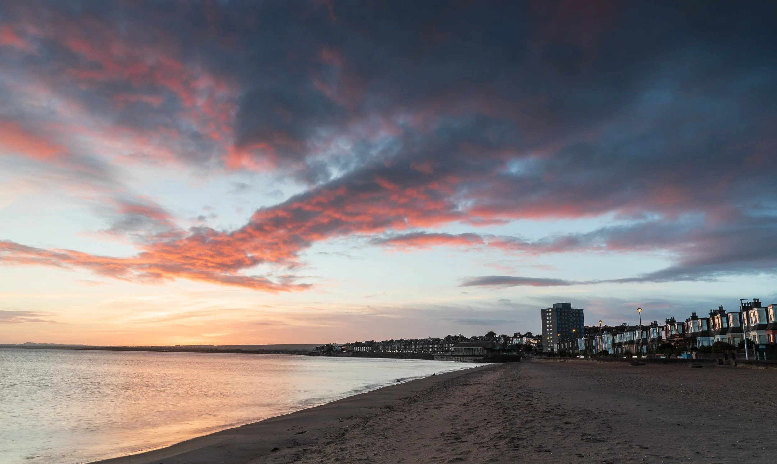 Sunset over a beach with calm water, sandy shoreline, and buildings in the distance, under a sky with pink and dark clouds.