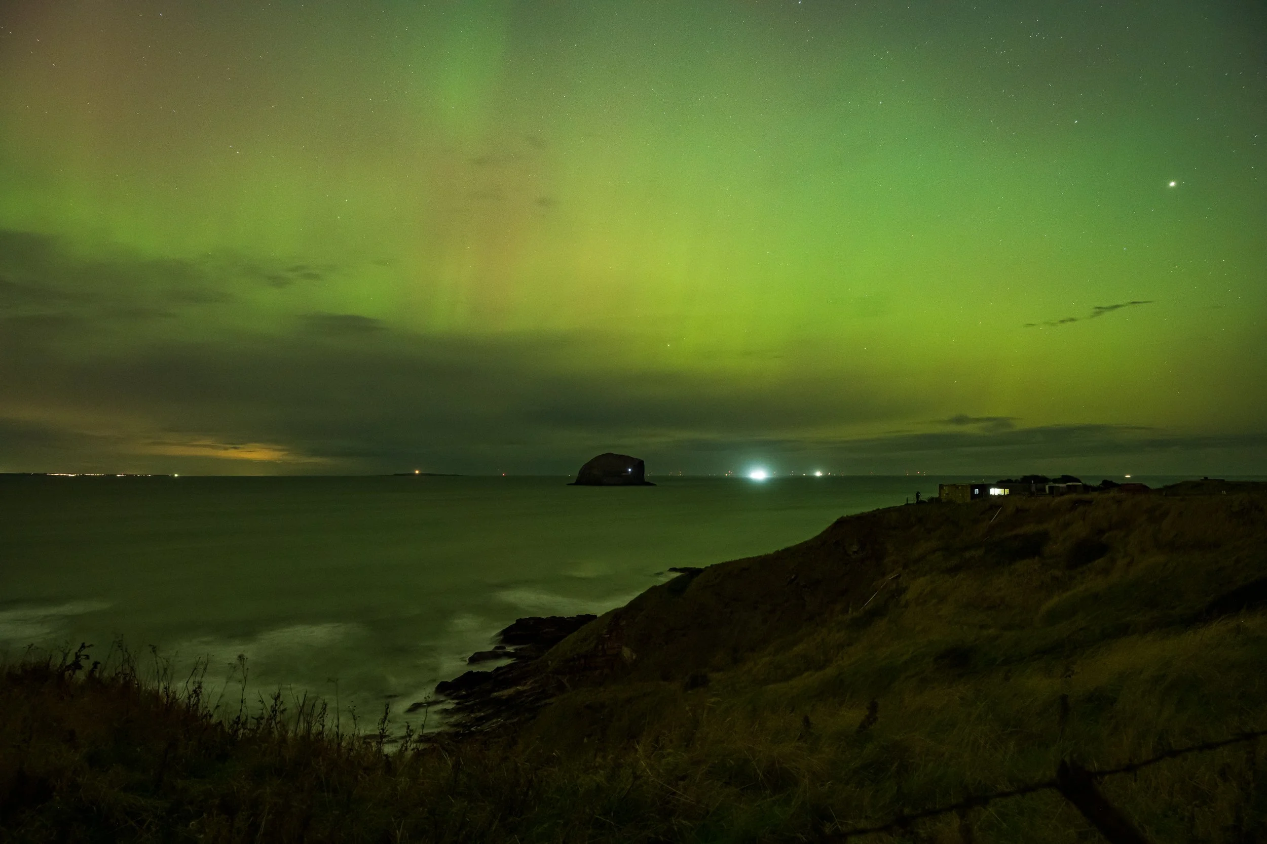 Nighttime view of the ocean with the Northern Lights in the sky and a large rock formation in the distance near the horizon. A grassy cliff is in the foreground with some small buildings or structures.