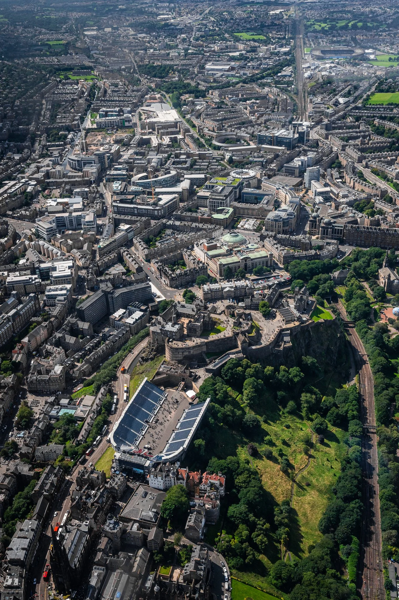 An aerial view of Edinburgh, Scotland, highlighting Edinburgh Castle atop Castle Rock, surrounded by city buildings and greenery, with nearby roads and railways.