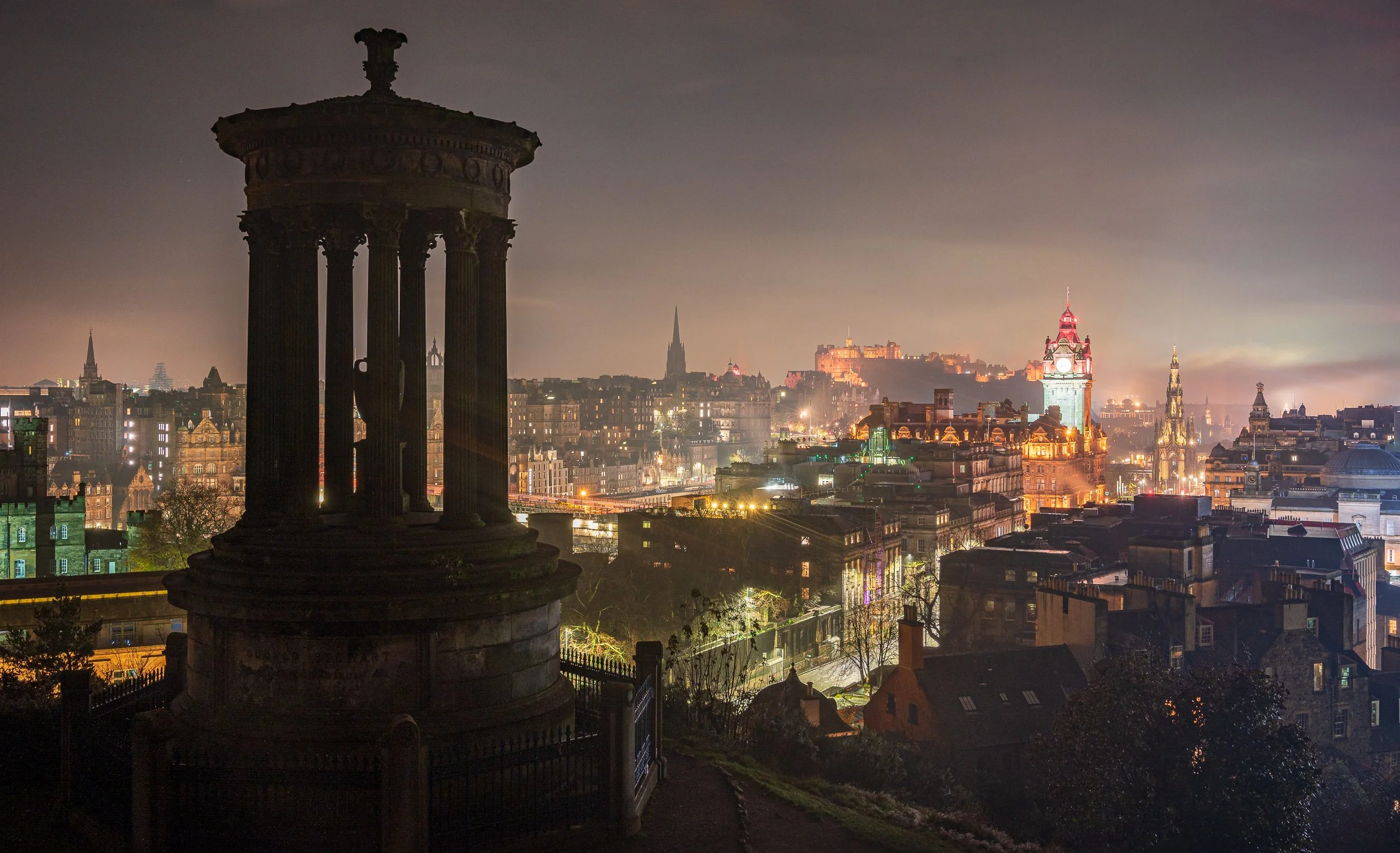 Nighttime cityscape of Edinburgh, Scotland, with illuminated historic buildings and a dark silhouette of a stone monument in the foreground.