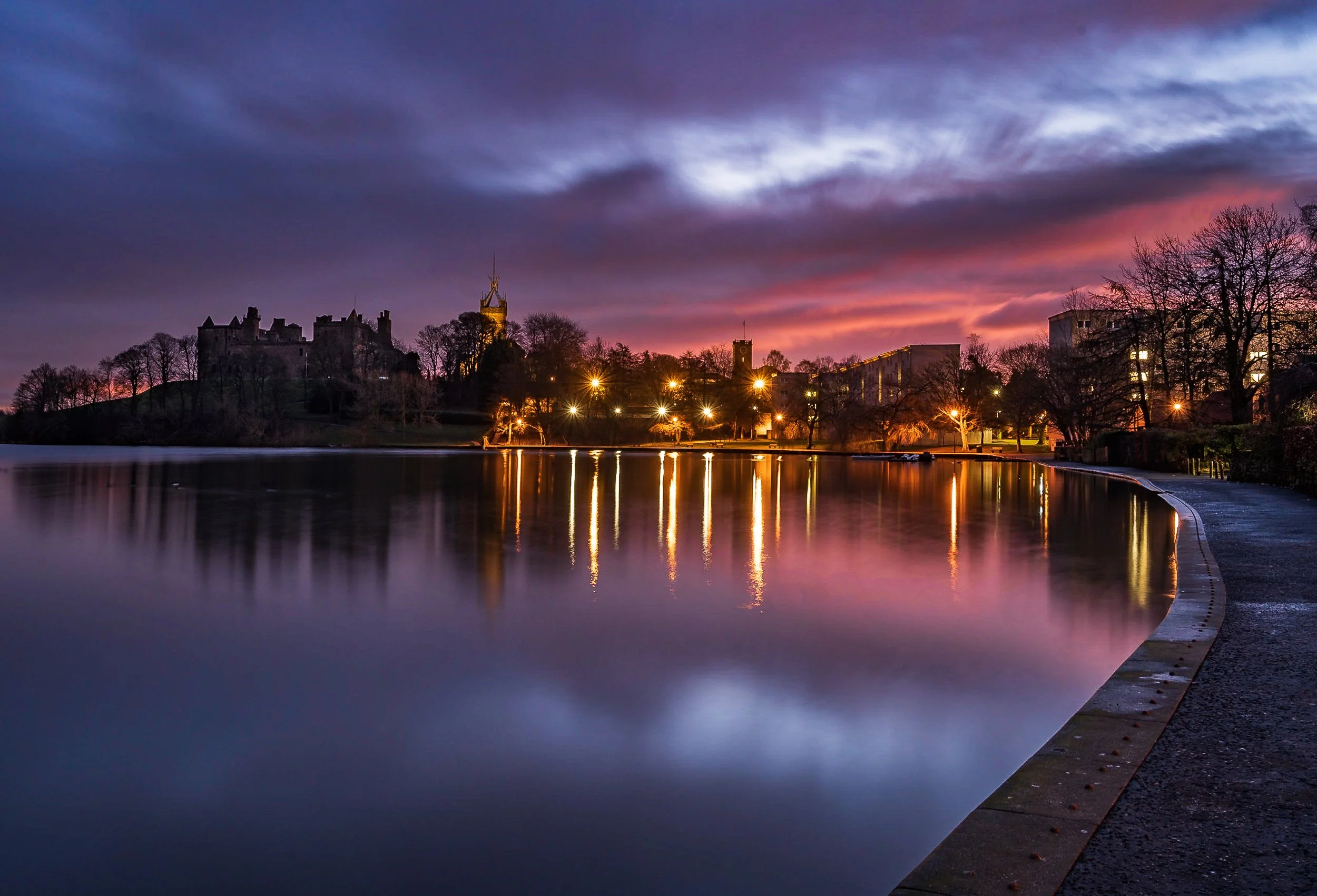 A twilight scene of a lake with reflections of lit streetlights, a castle on a hill, and buildings against a purple and pink sky with clouds.
