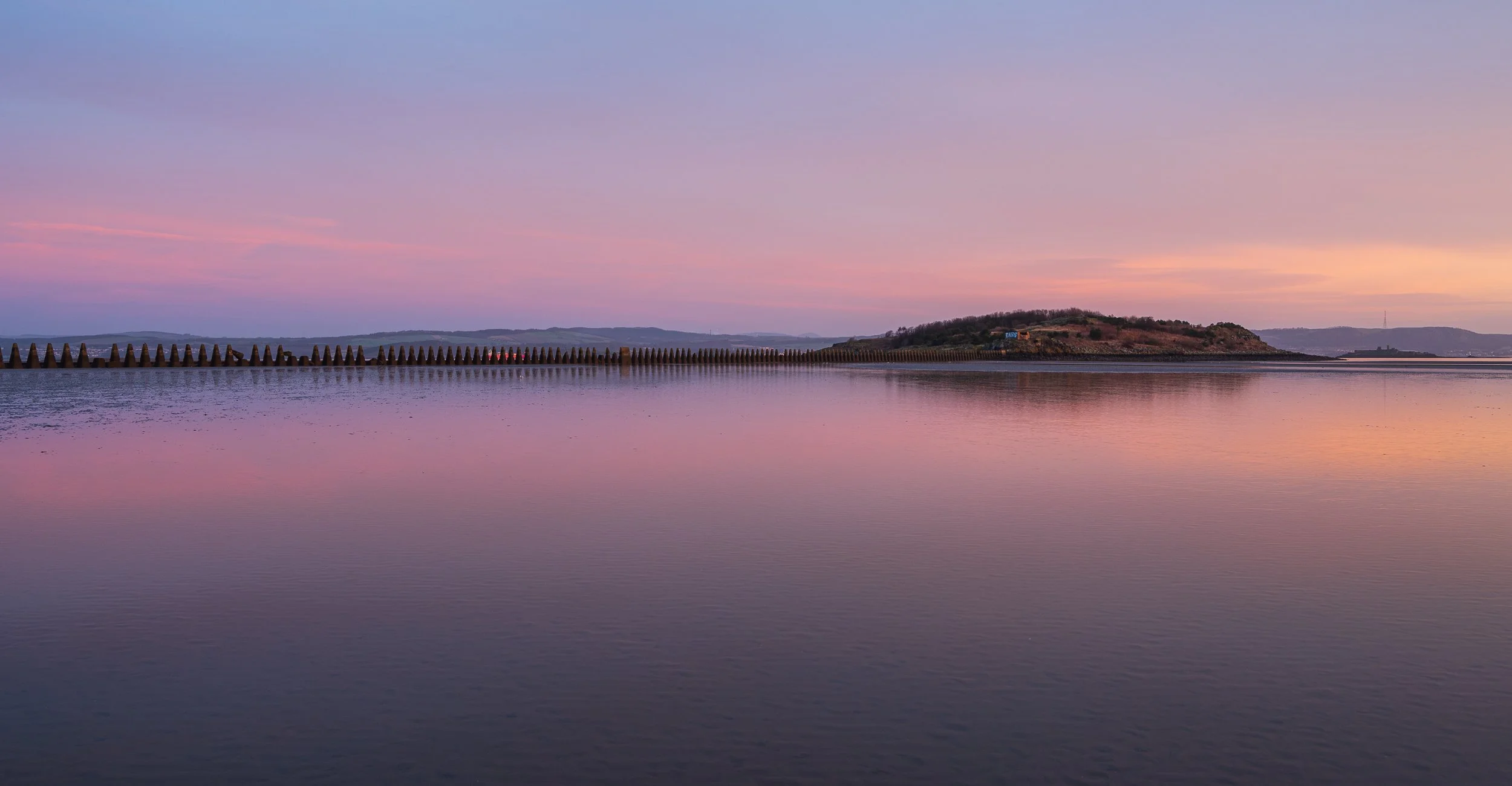 A peaceful lakeside scene at sunset with pink and purple sky, calm water reflecting the colors, a small island, and a long breakwater with cone-shaped structures.