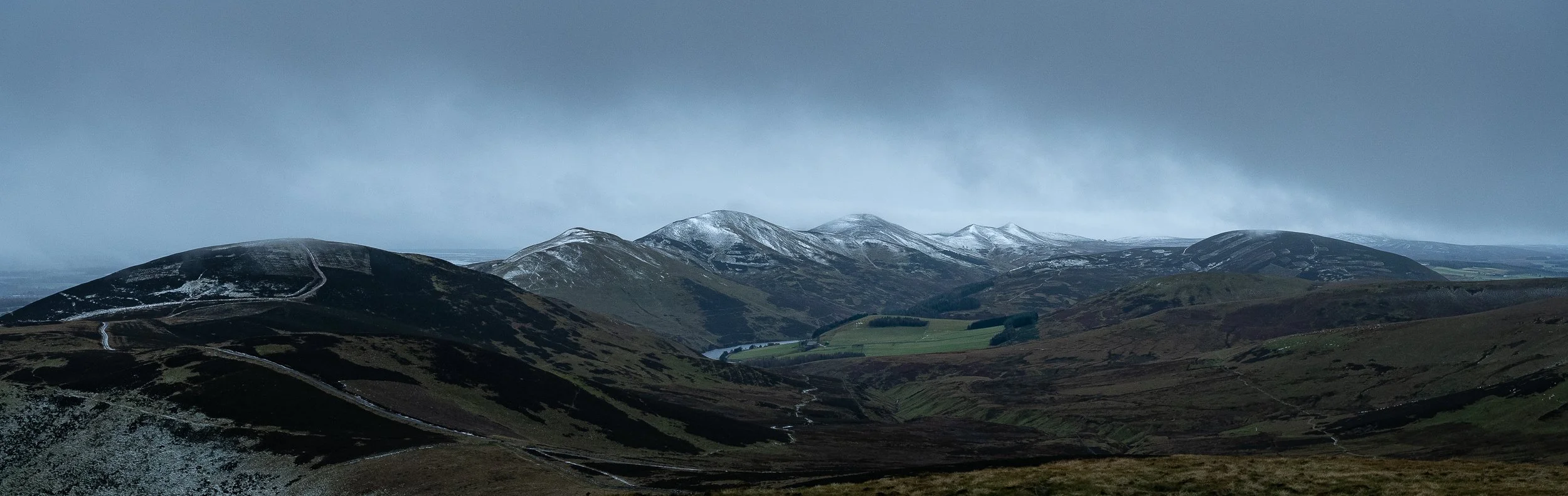 Landscape of rolling hills and mountains with patches of snow on the peaks under a cloudy sky.