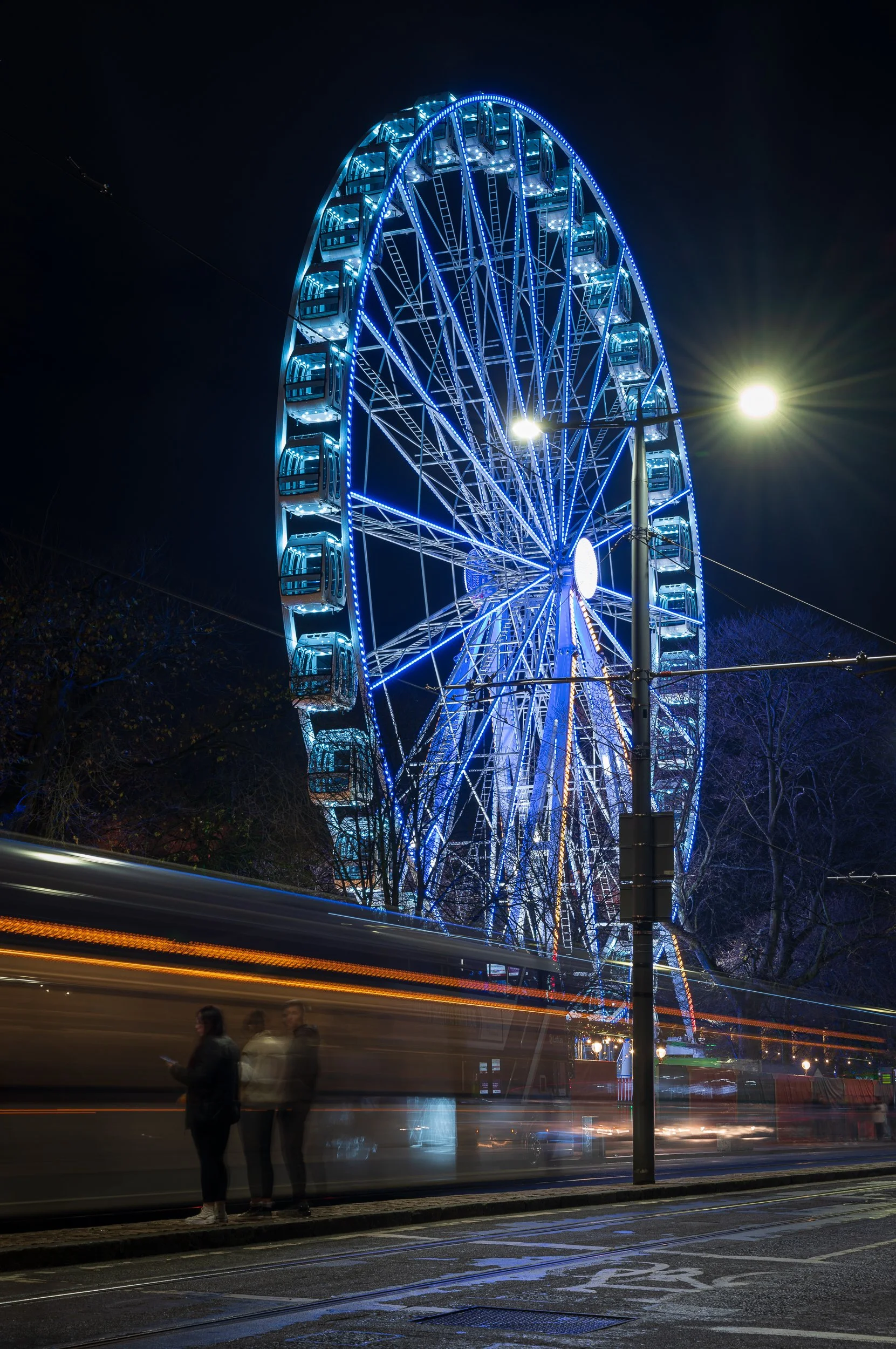 Night scene of a brightly lit amusement park Ferris wheel with blue neon lights, triple motion-blurred pedestrians and tram in the foreground, streetlights, and dark sky.