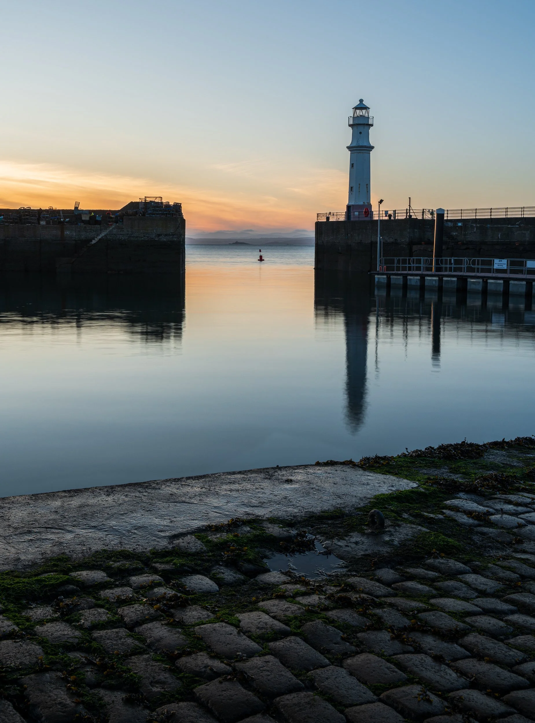 A lighthouse on a pier at sunset with calm water and rocky foreground.