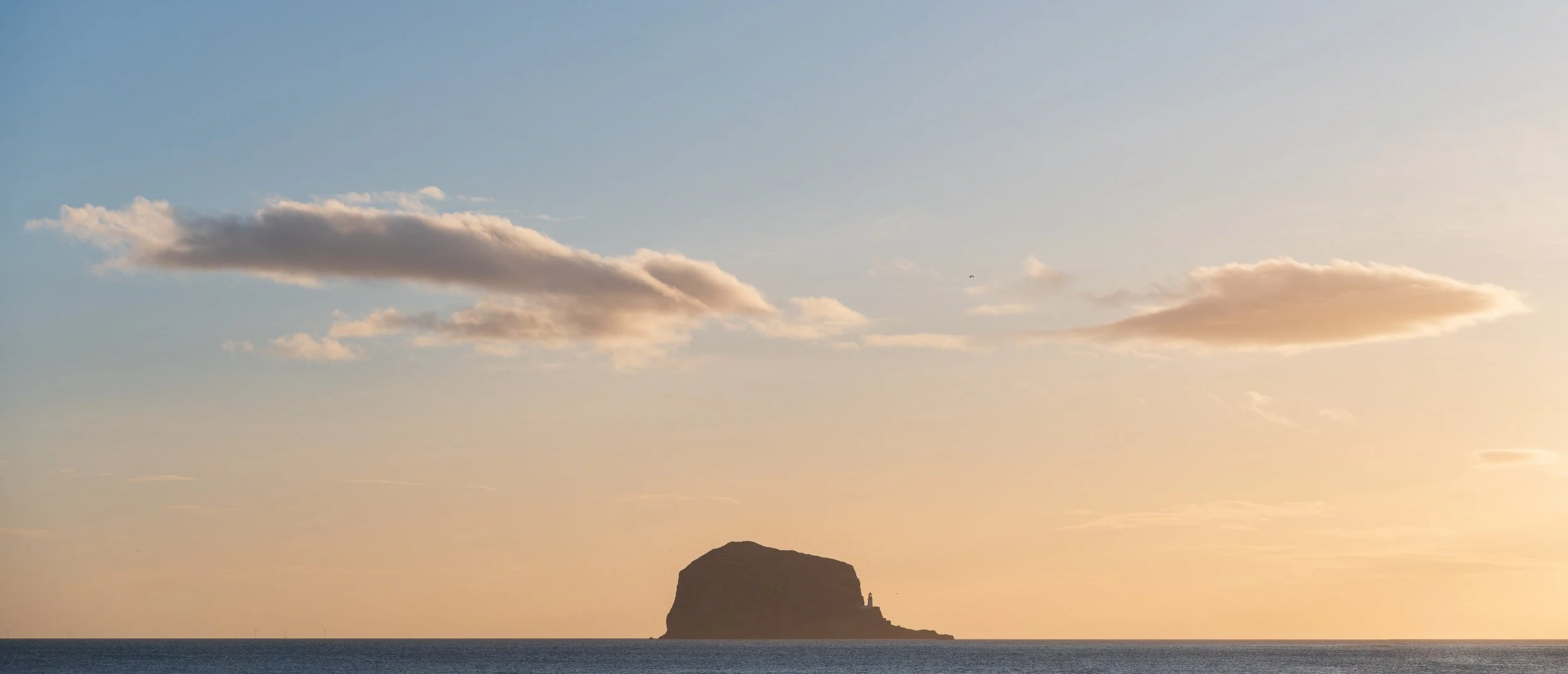 Sunset over the ocean with a rocky island and a lighthouse silhouette, two clouds in the sky.