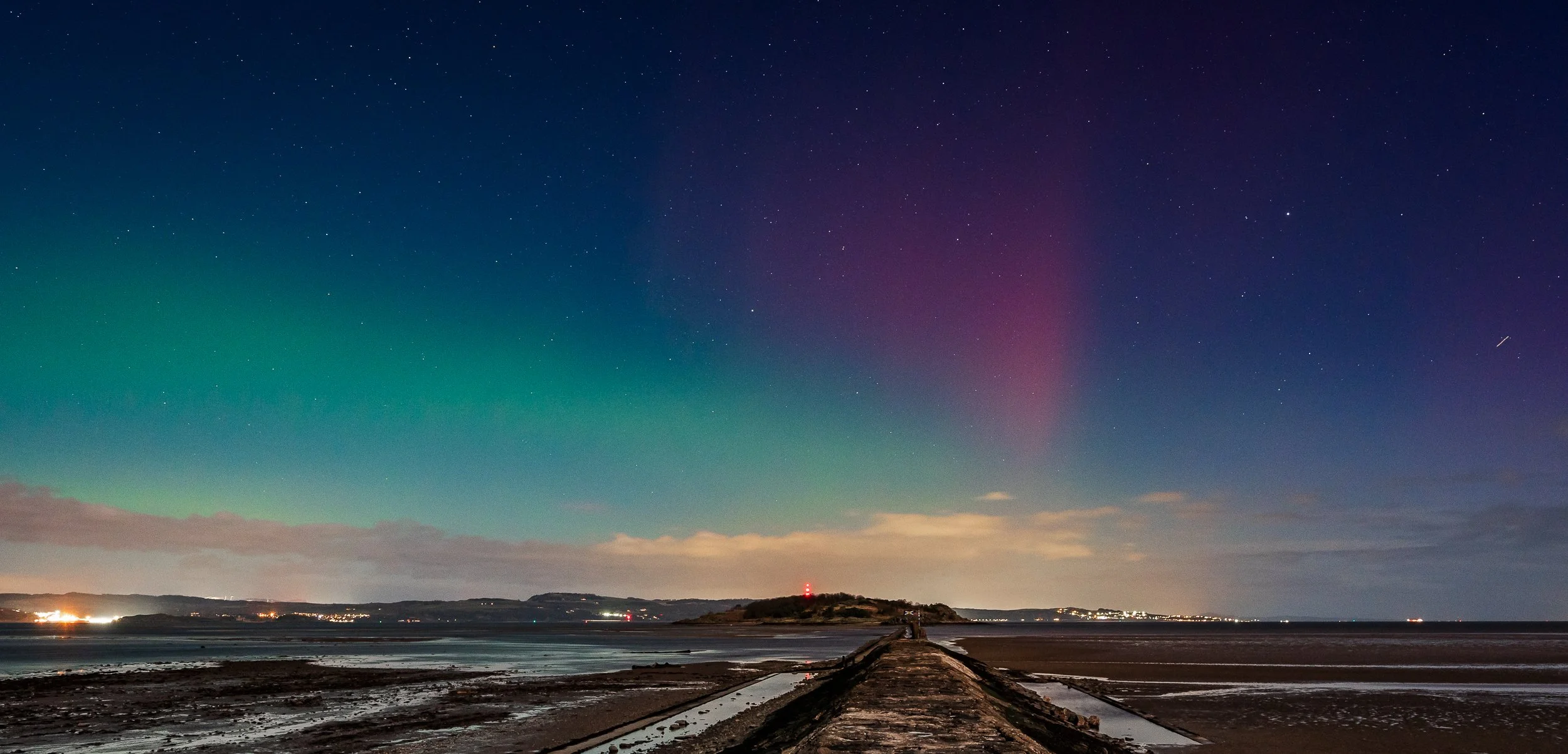 Nighttime view of the northern lights over a coastal landscape with a pier leading to an island, stars visible in the sky.