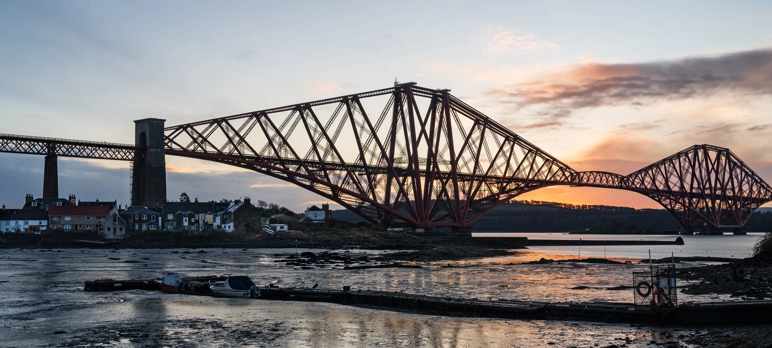 A large red and black iron bridge spans across a river at sunset, with small houses along the shore and boats docked in the foreground.