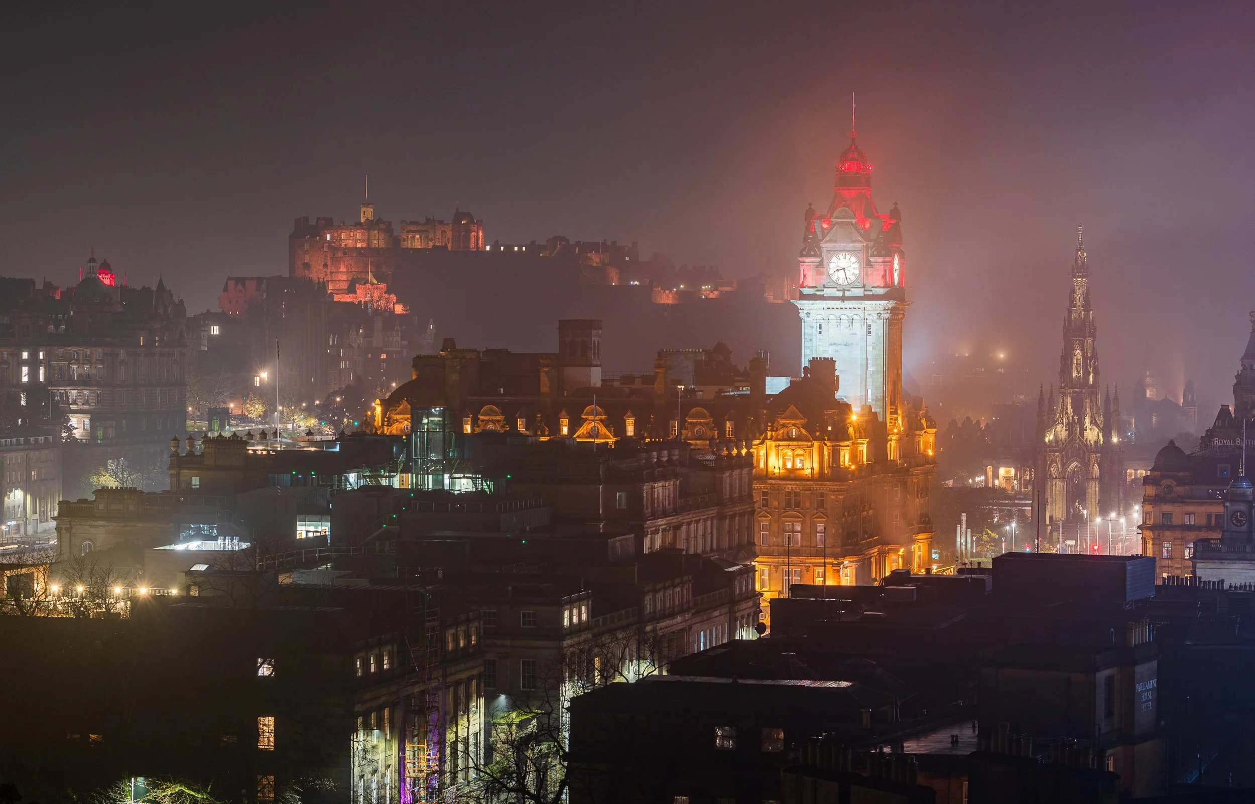 Night view of Edinburgh city with illuminated buildings, historic clock tower, and Edinburgh Castle on a hill, all shrouded in fog.