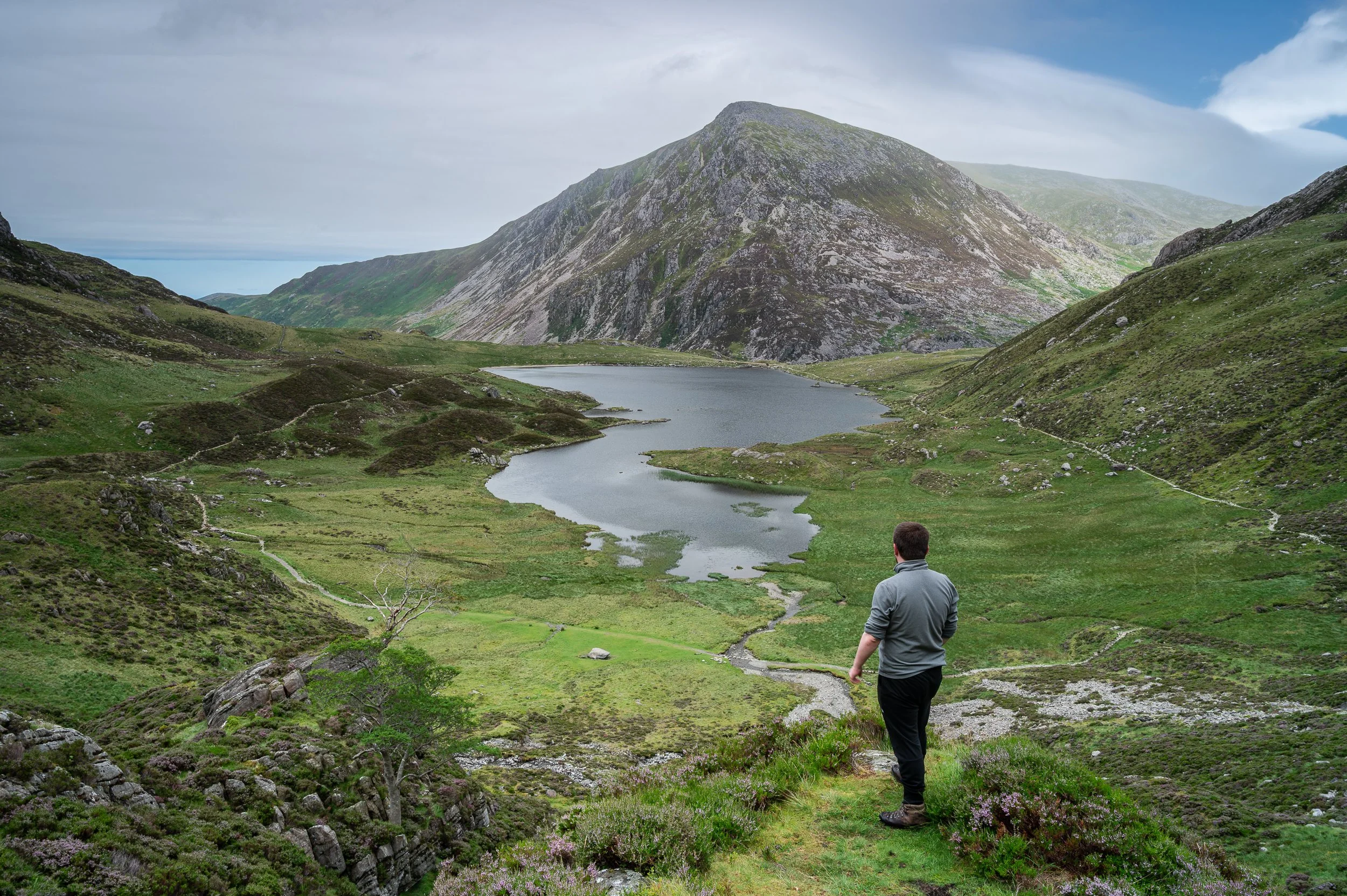 Man in gray jacket and black pants standing on a grassy trail overlooking a lake in a mountainous landscape with green hills and a cloudy sky.