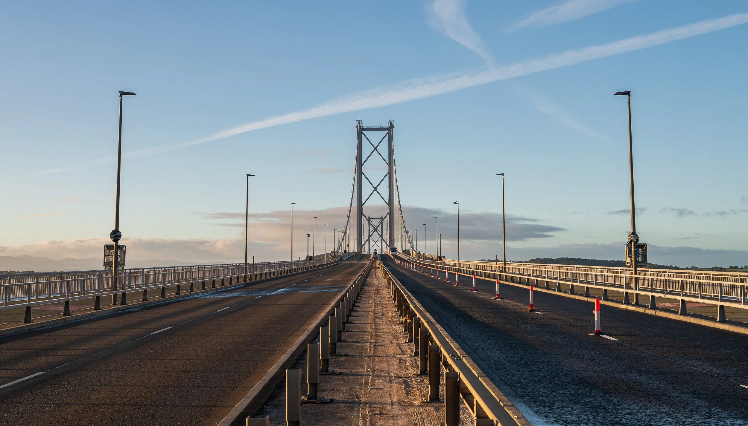 Empty bridge with streetlights and railings, spanning a body of water, under a blue sky with clouds and jet trails.