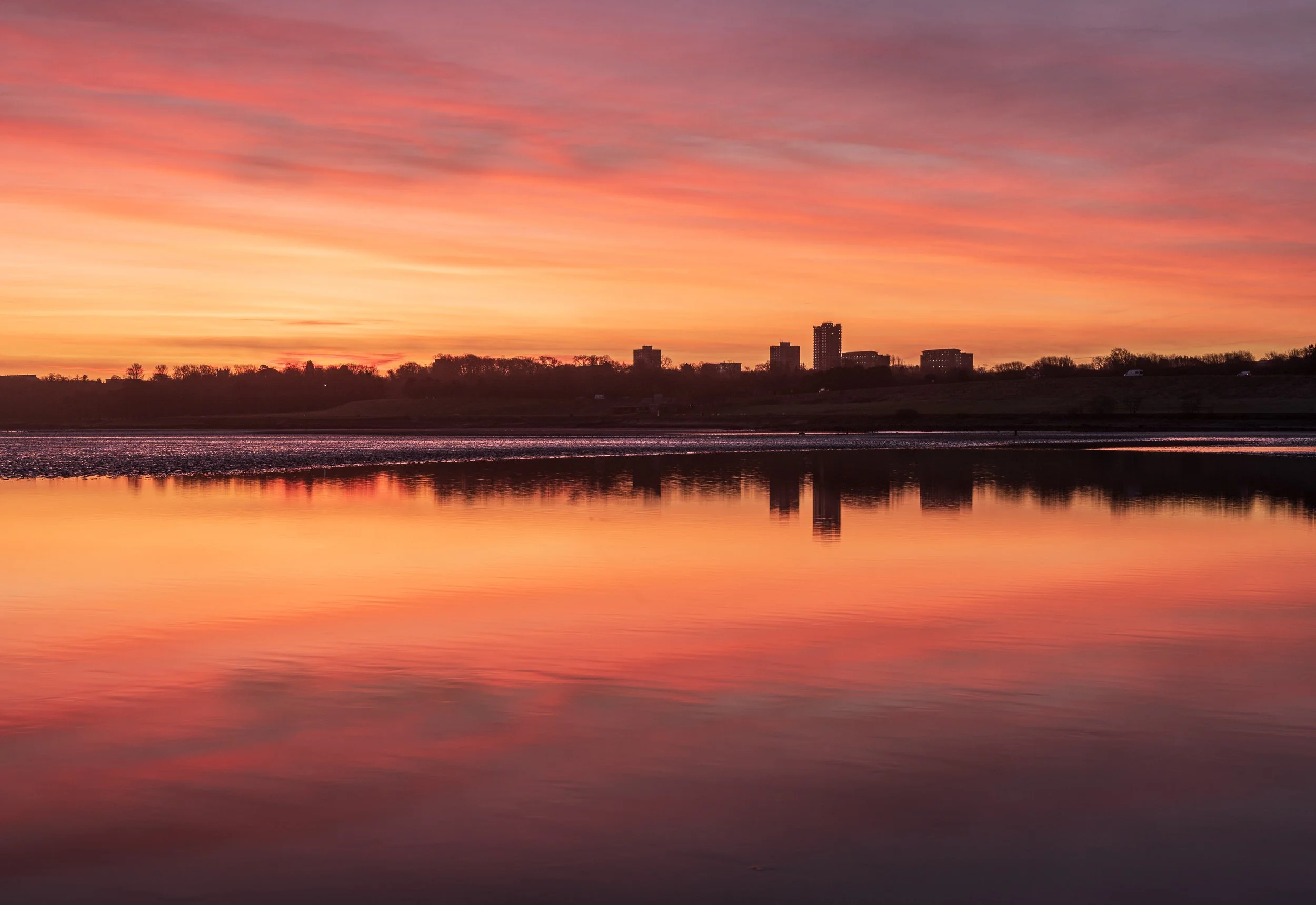 Sunset over a body of water reflecting the orange and pink hues of the sky, with a distant city skyline silhouette on the horizon.
