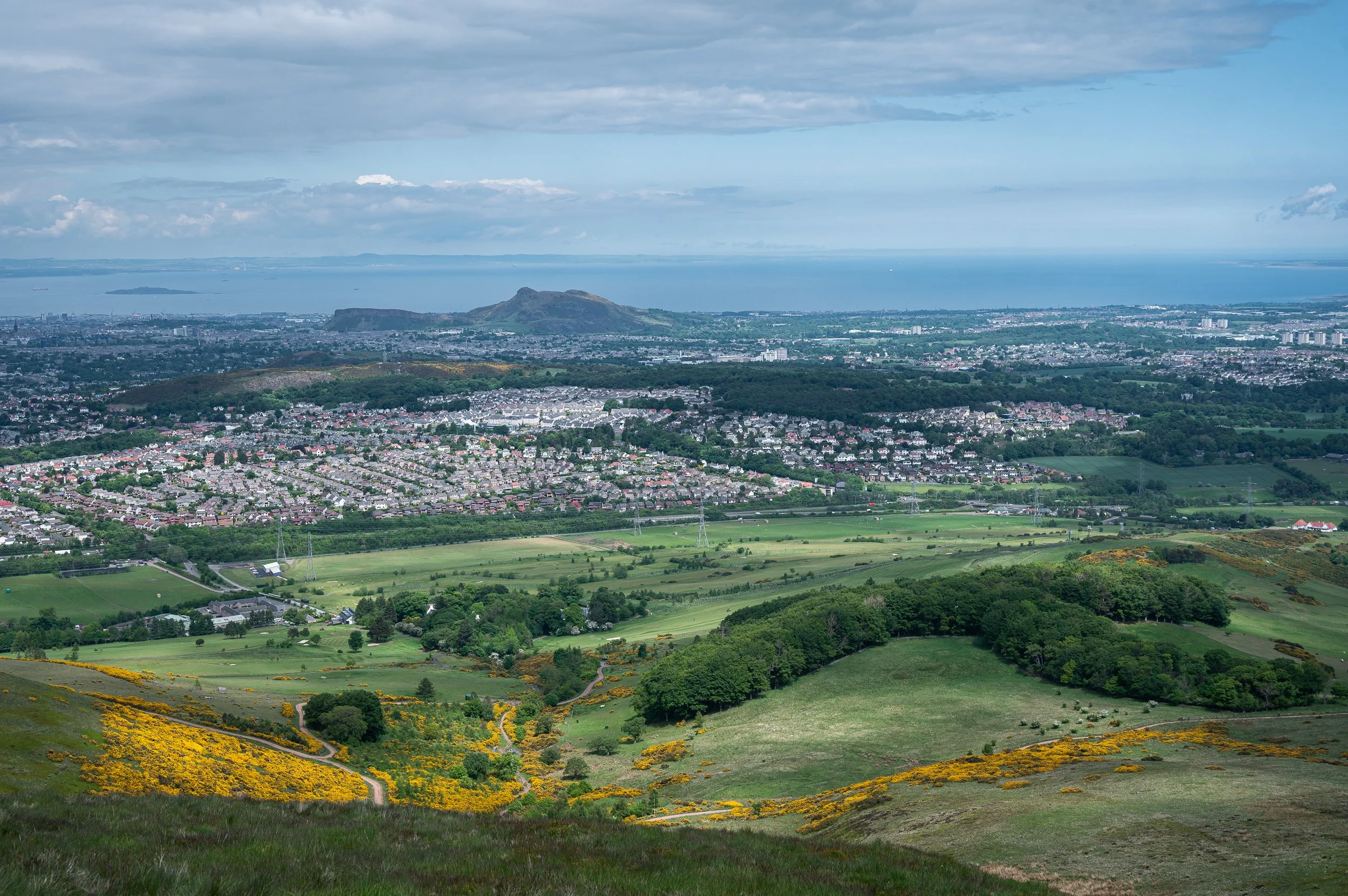 Scenic view of a city and surrounding green hills with yellow flowers, fields, and forests, overlooking water and distant islands under a partly cloudy sky.