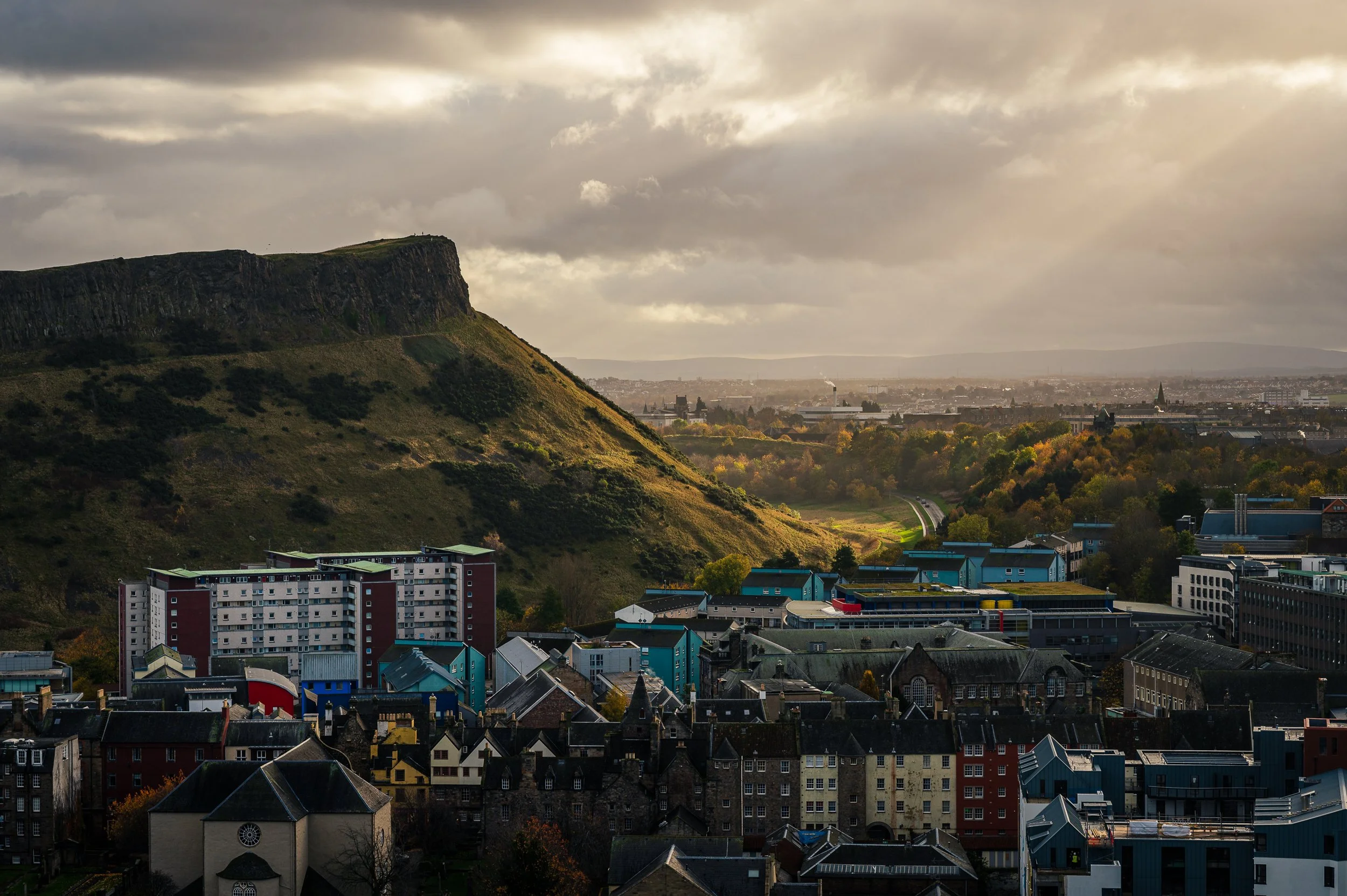 Cityscape with colorful buildings, green hills, and a mountain with a flat top in the background under a cloudy sky.