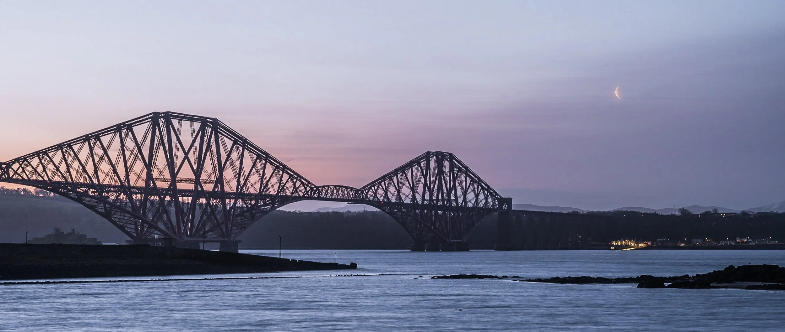 Silhouette of a large steel bridge over water during dusk, with a crescent moon in the sky and a distant shoreline with city lights in the background.