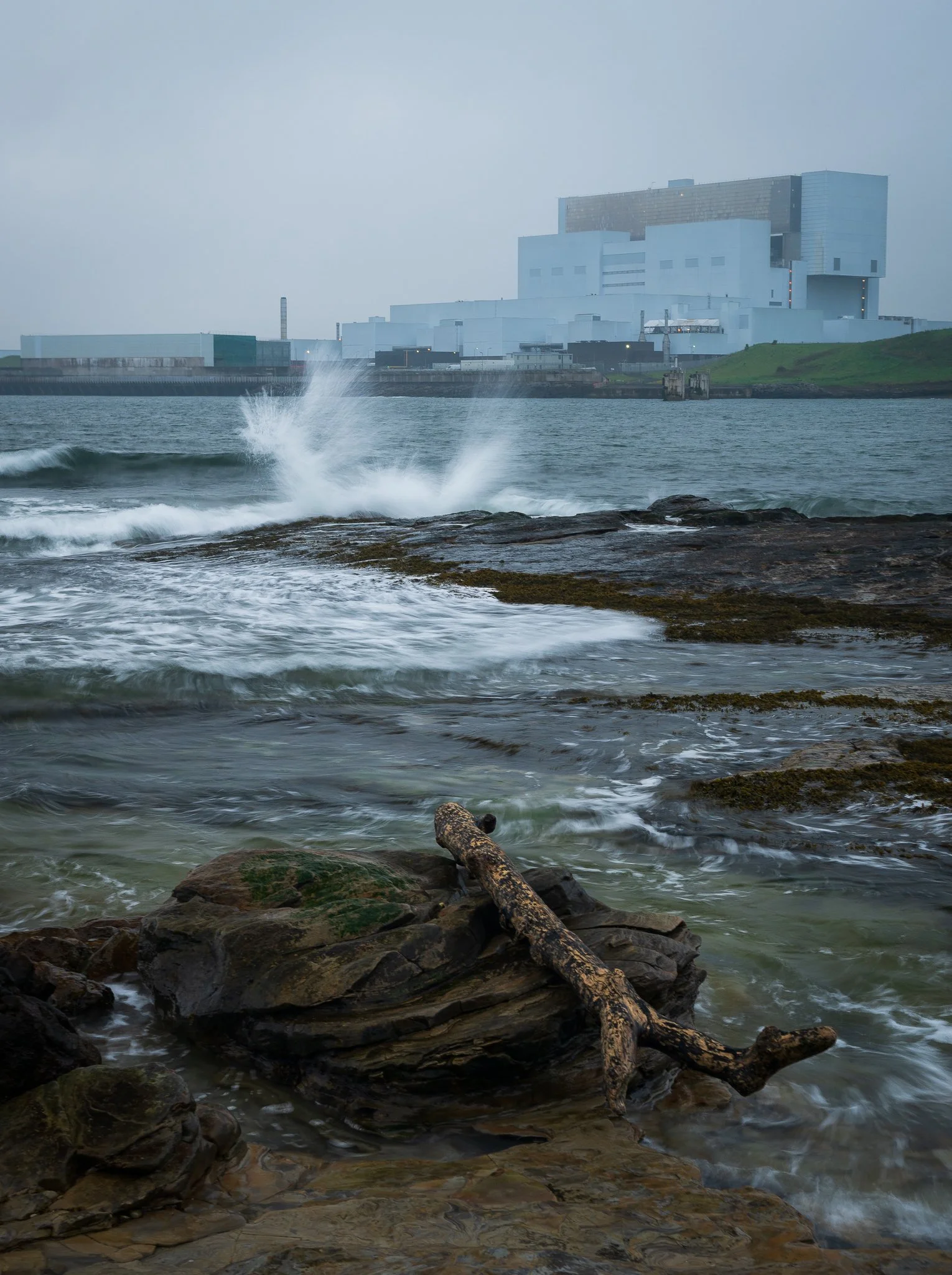A rocky shoreline with weathered driftwood in the foreground, ocean waves crashing against the rocks, a large industrial building in the background, and a cloudy sky.