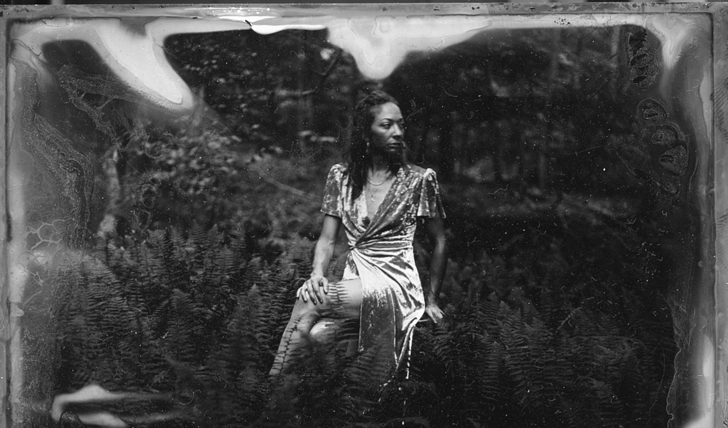 A black and white photograph of a woman sitting outdoors in a natural setting. She appears to be deep in thought, wearing a patterned dress, with her hand resting on her knee. The background consists of dense foliage, and the image has a vintage, slightly worn look with visible scratches and imperfections.