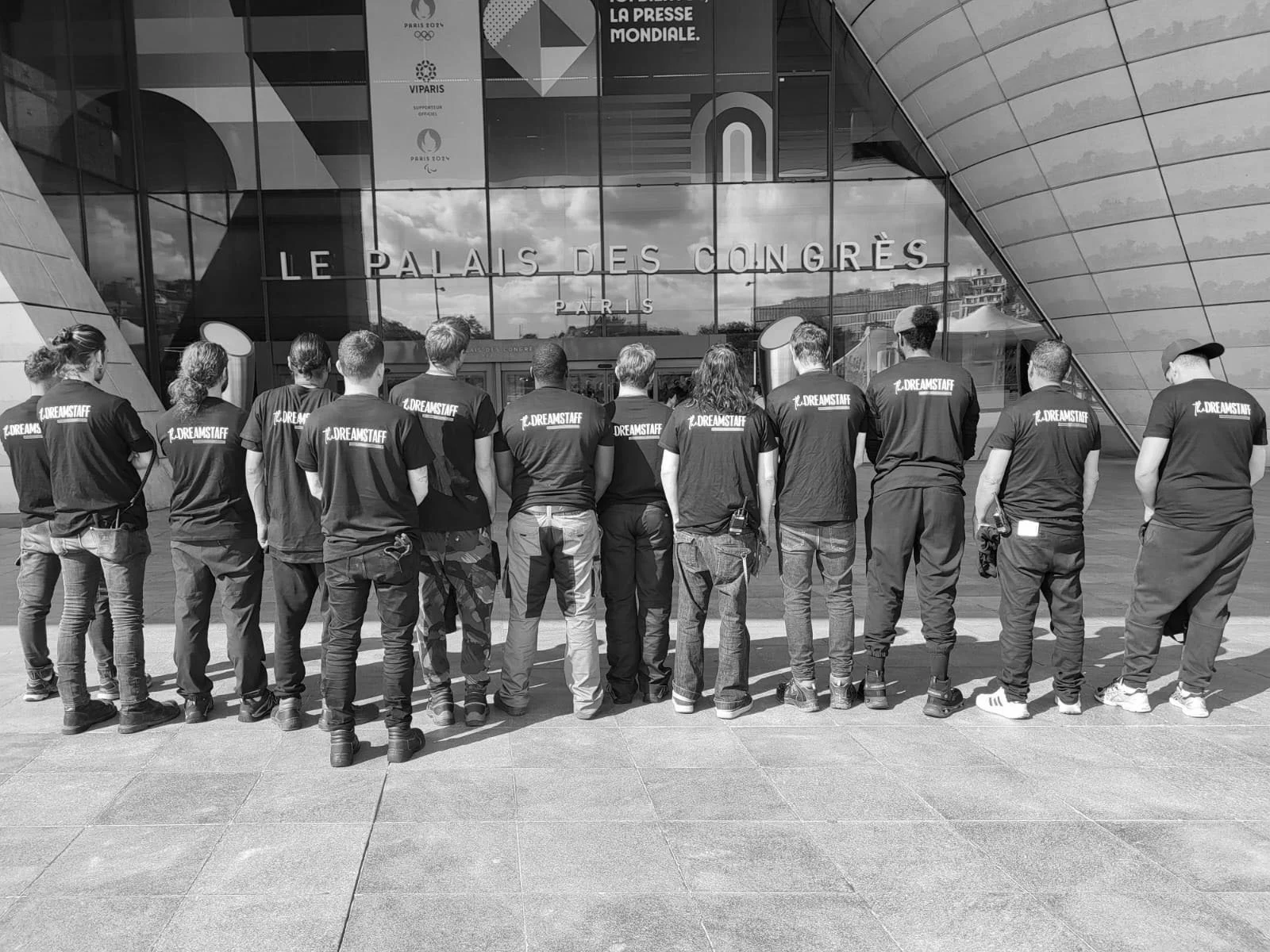 Groupe de neuf jeunes personnes debout sur une surface asphaltée, certaines portant des t-shirts avec inscription 'MINI DANCING CLUB', photographiées en noir et blanc.