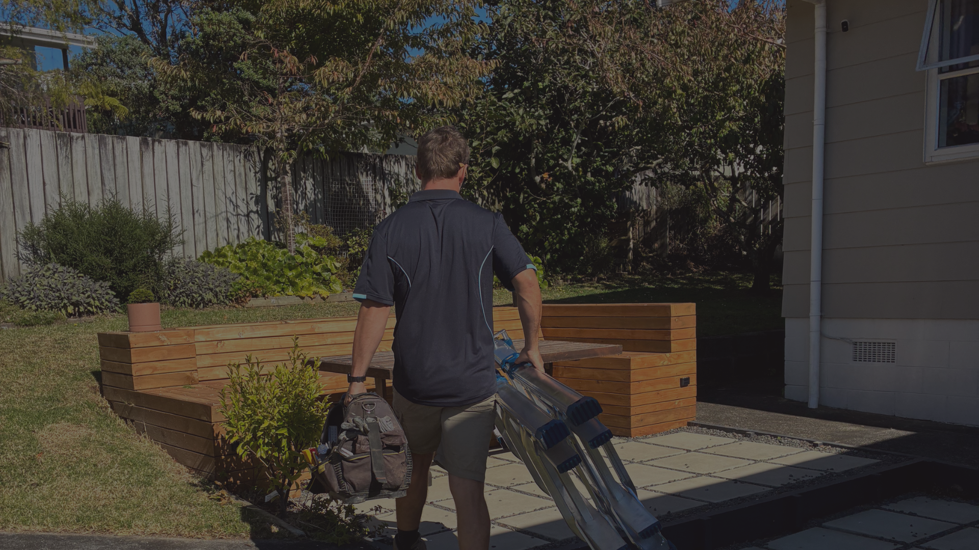 A man with a backpack walking away while carrying a rolled-up metal framework in a backyard patio with wooden benches, plants, and a fence and trees in the background.
