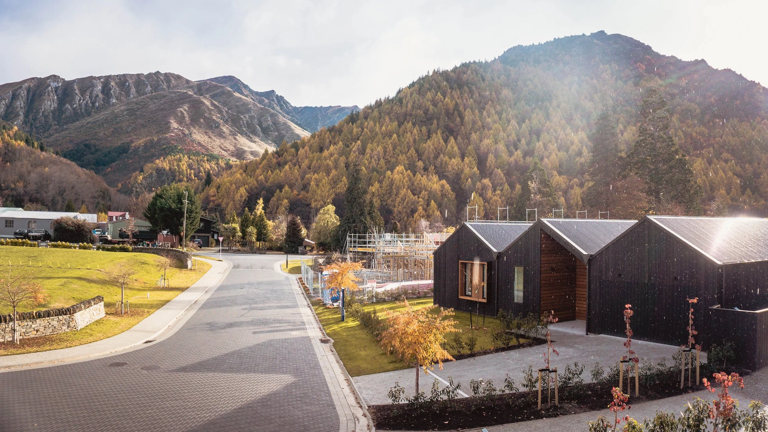 A mountain landscape with a small neighborhood of modern dark houses and a winding paved road in the foreground, surrounded by trees and autumn foliage.