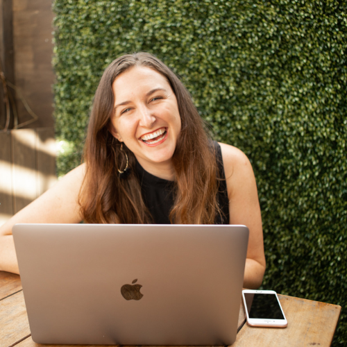 Smiling woman with long brown hair wearing a black sleeveless top, sitting at a wooden table with a silver MacBook and a smartphone outdoors, with a green hedge background.