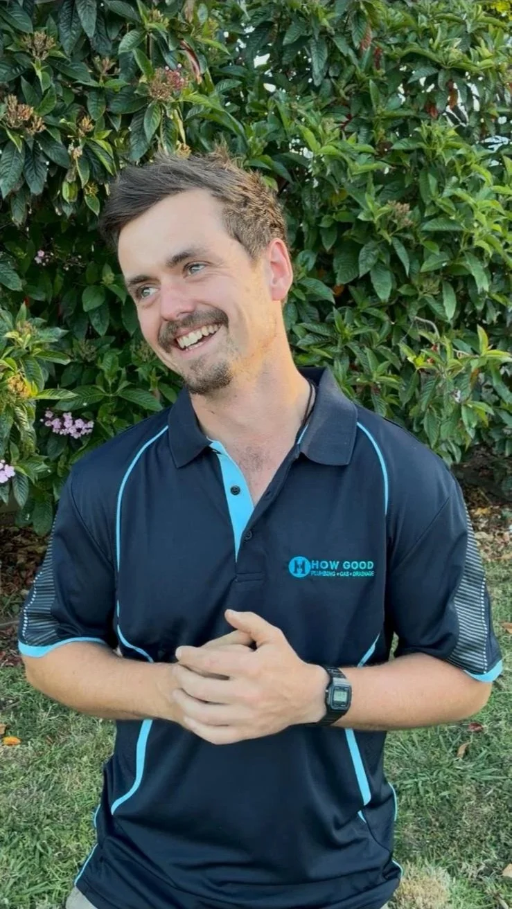A smiling man with short dark hair and a beard, wearing a navy blue sports shirt with light blue accents, standing outdoors in front of green foliage and small purple flowers. The shirt has a logo that says 'HOW GOOD Plumbing Gas Drains.'