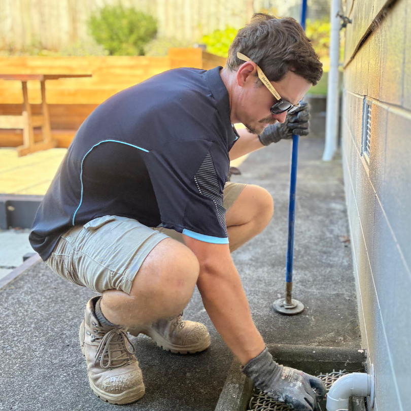 A man kneeling outside, working on a drain or pipe near a building wall with a blue tool, wearing gloves, sunglasses, a black sports shirt, and khaki shorts.