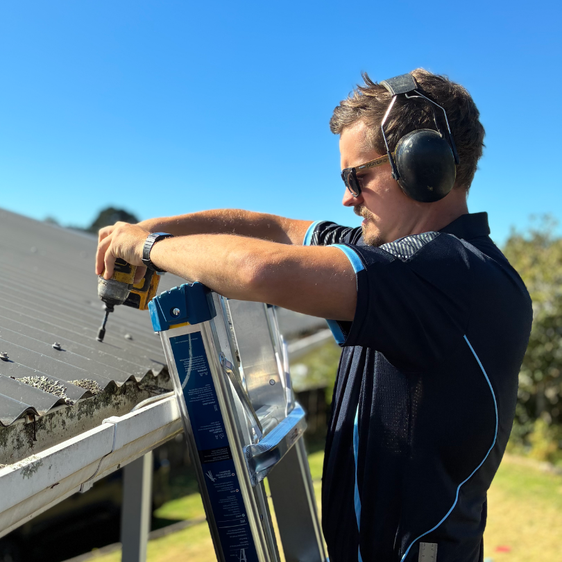 A man wearing safety headphones and sunglasses is using a power drill while standing on a ladder to work on a metal roof outside on a sunny day.
